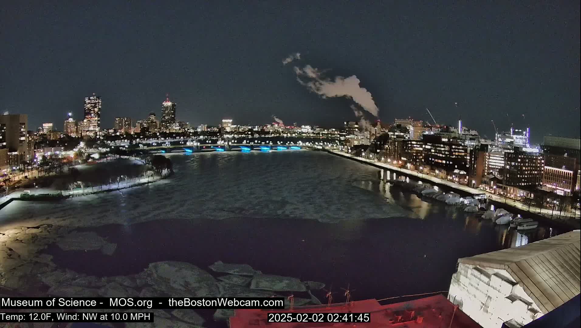 A nighttime cityscape with a partially frozen river, illuminated buildings, and smoke rising from a distant source, under a dark sky.