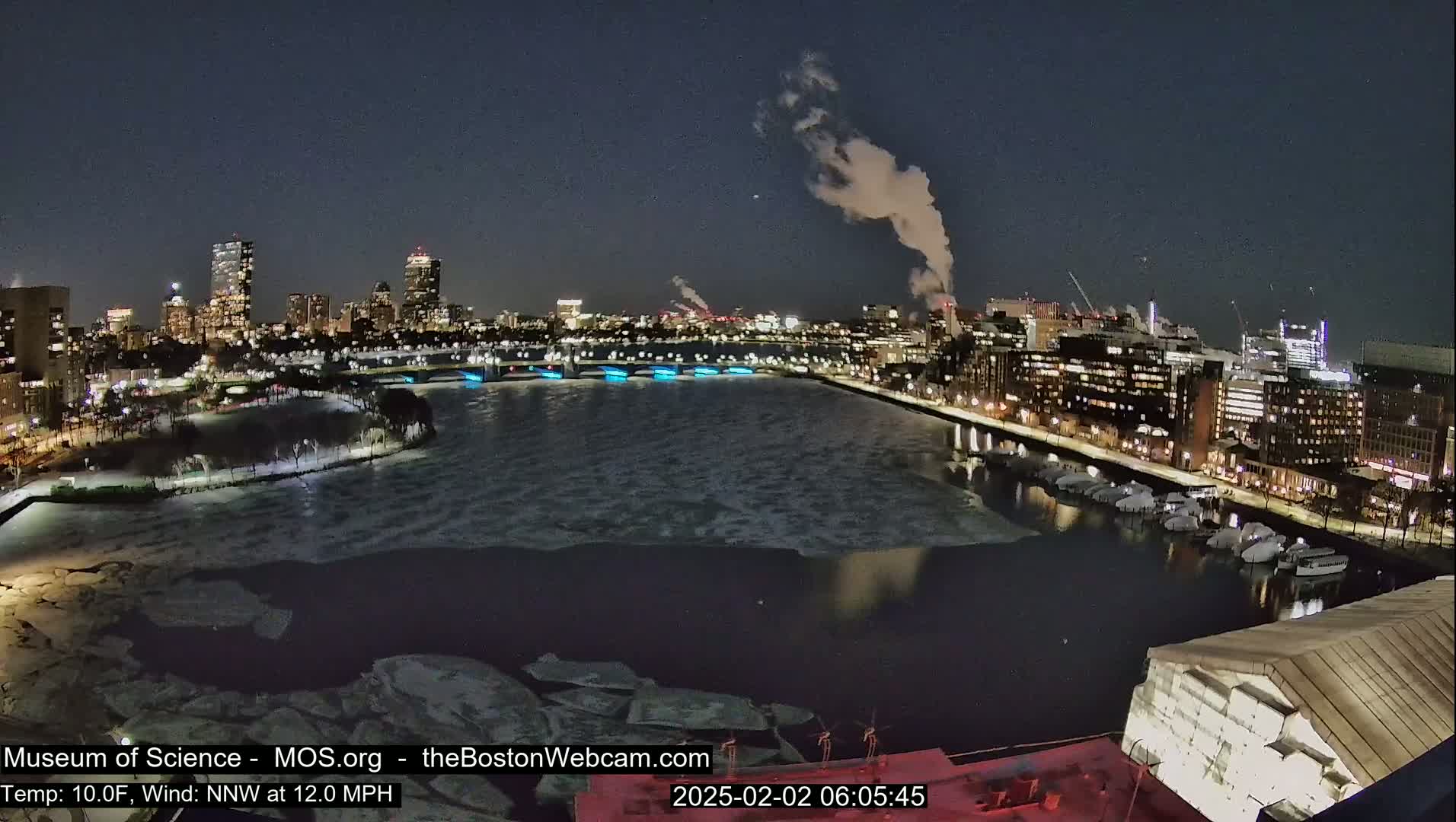 A nighttime view of a city skyline across a partially frozen river, with some smoke rising from a distant source and a 10°F temperature with a 12 mph north-northwest wind.