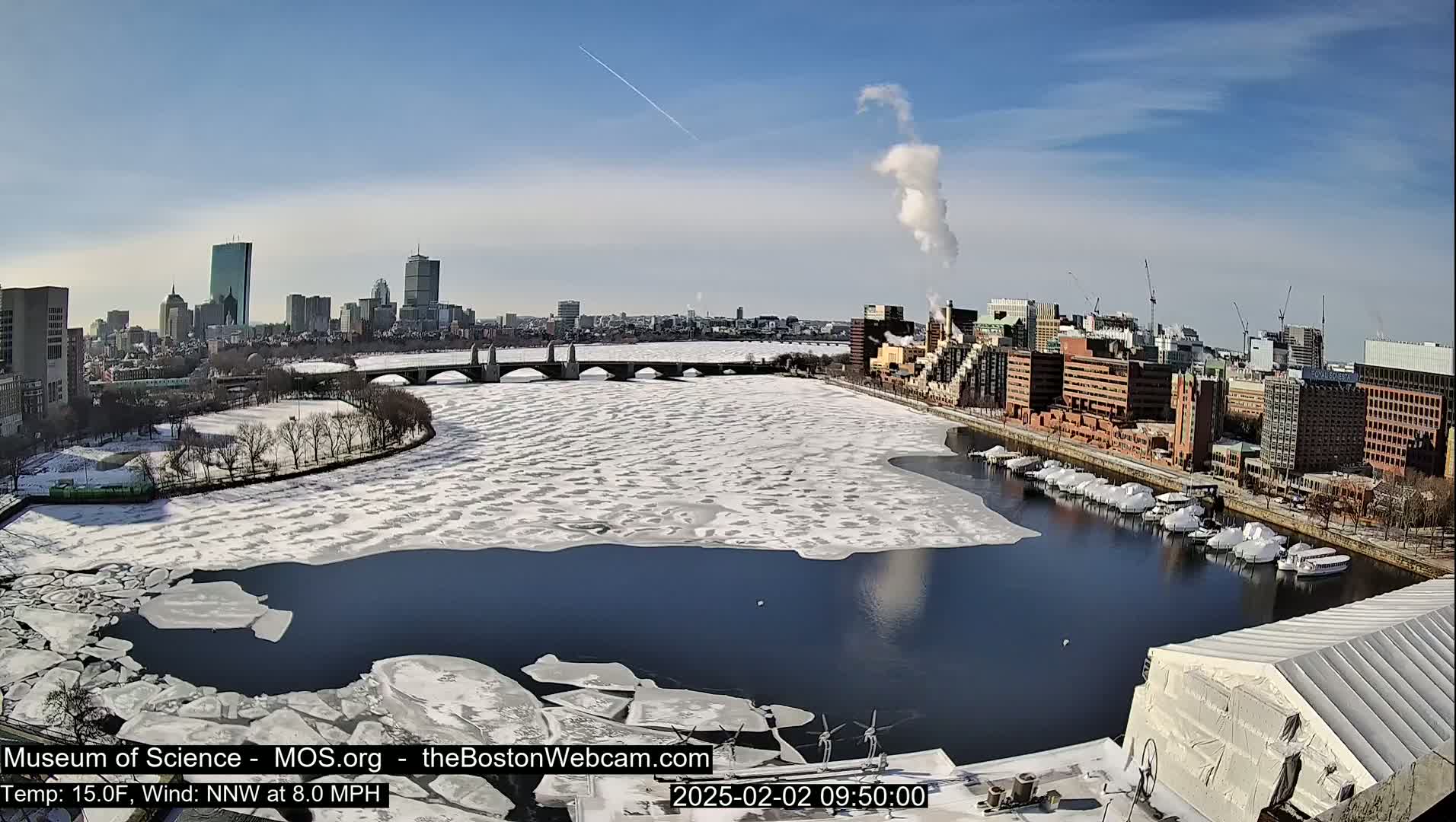 A partly frozen river flows through a city with a bridge and skyline under a clear, sunny sky.