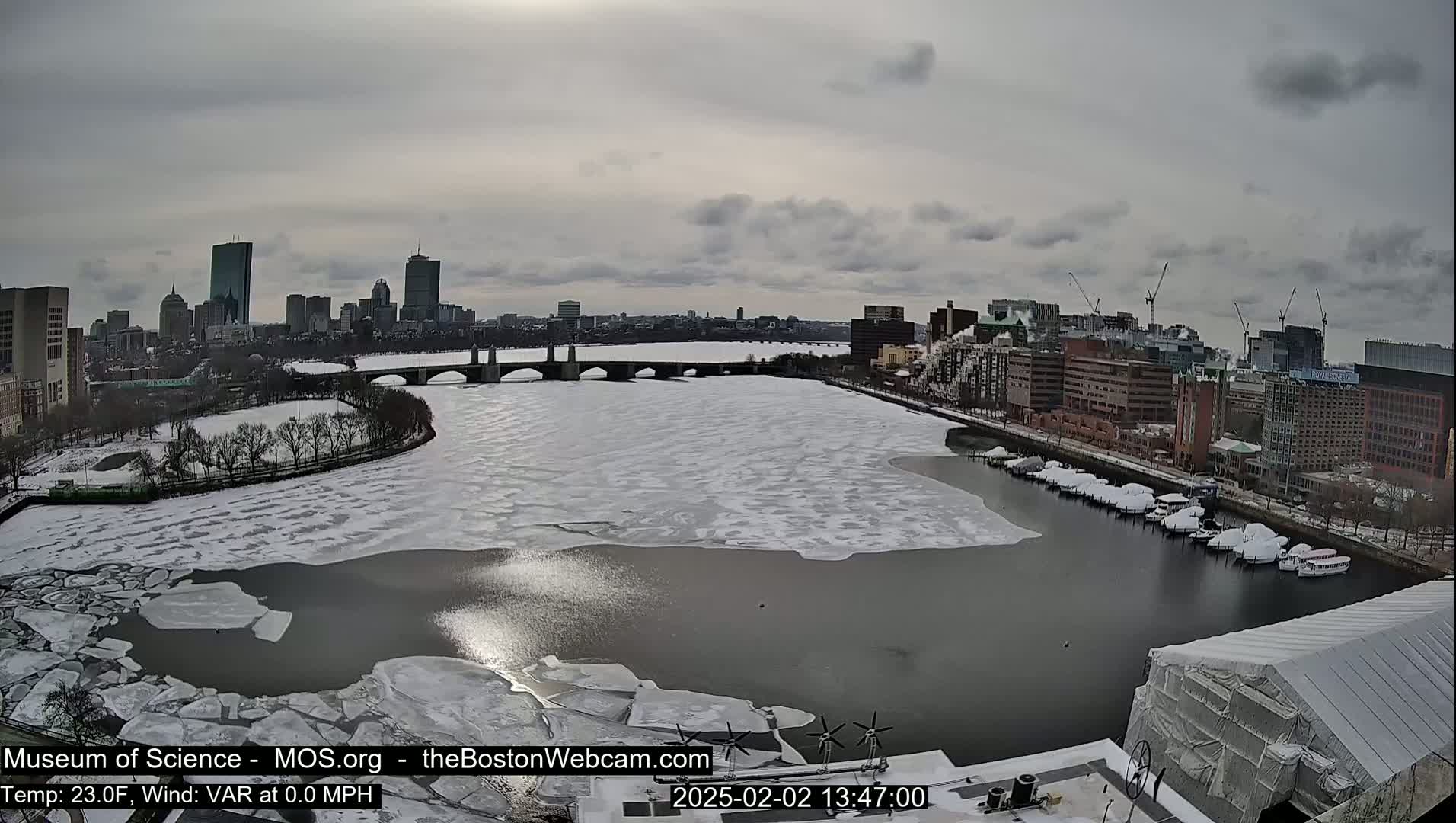 A partially frozen river flows through a city under a cloudy sky, with boats docked along the shore and a cityscape visible in the distance.