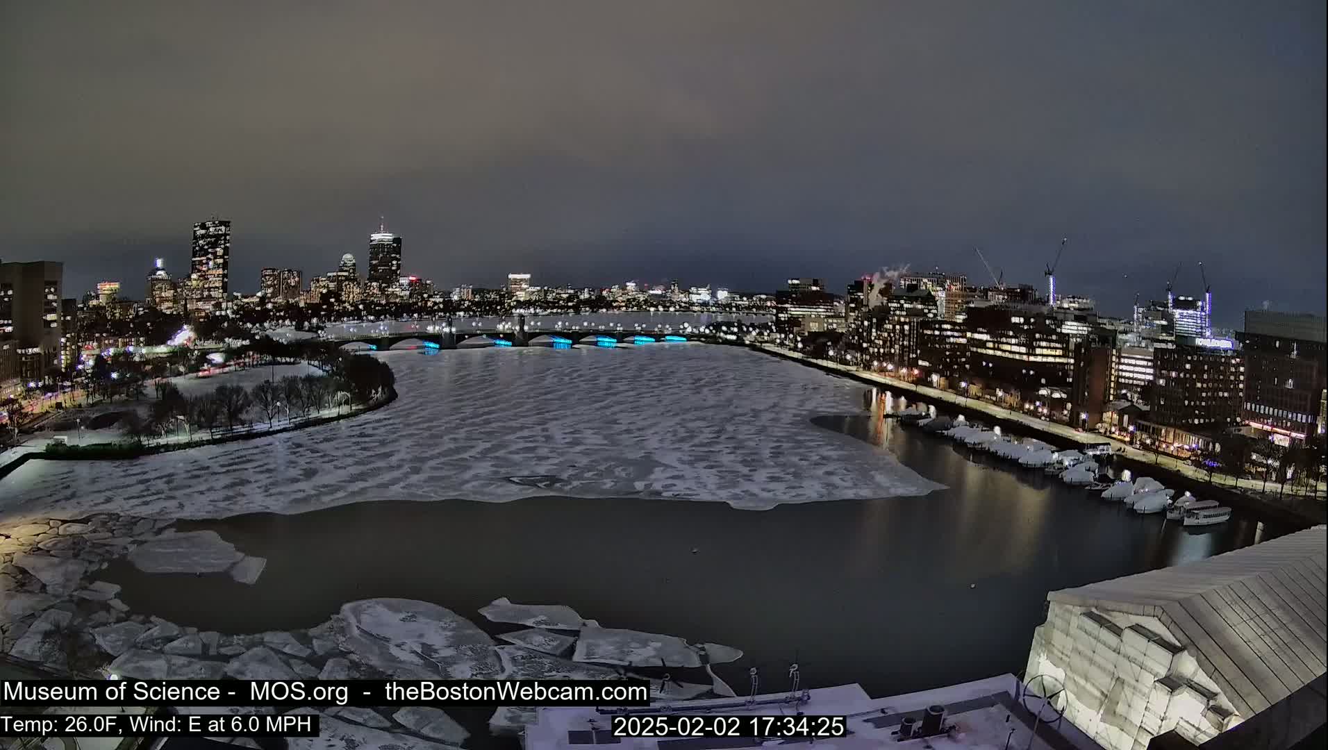 A nighttime cityscape with a partially ice-covered river, illuminated bridges, and docked boats under a dark sky.