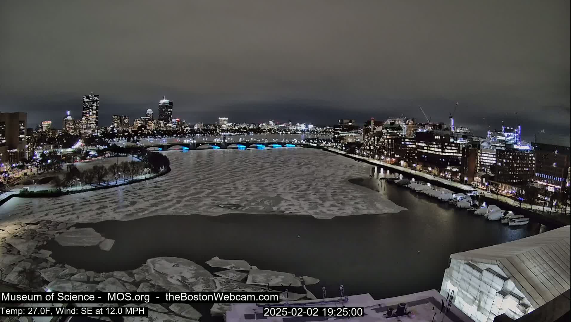A nighttime cityscape with a partially frozen river, illuminated bridges, and docked boats under a dark, overcast sky.