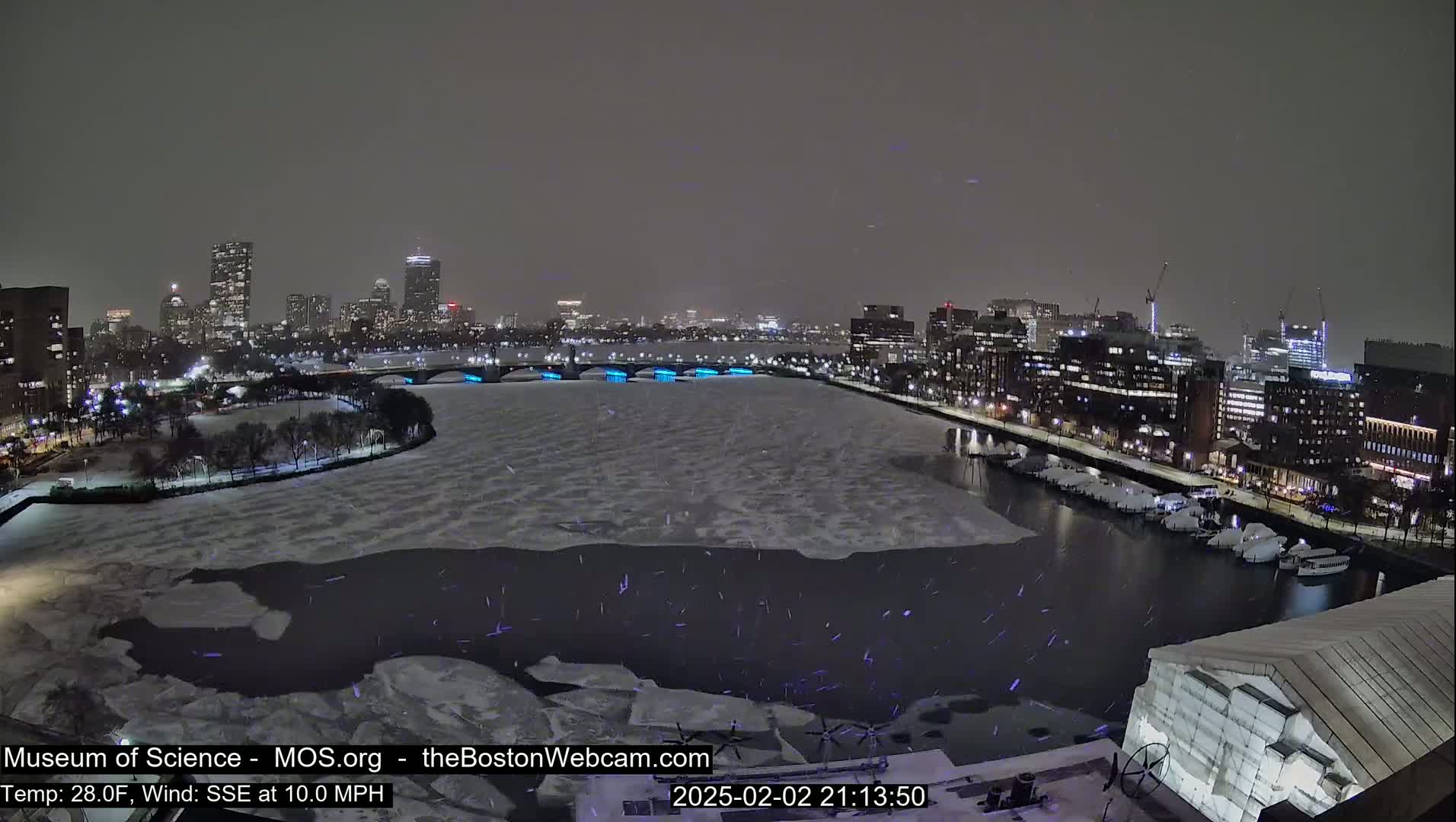 A snow-covered cityscape at night, with a partially frozen river in the foreground, is illuminated by city lights under a dark sky.