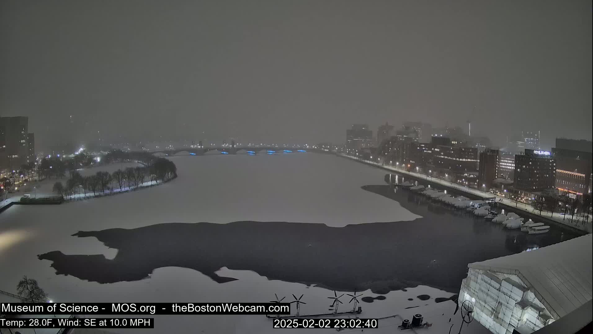 A snow-covered cityscape at night, viewed from above, shows a partially frozen river with buildings and a bridge illuminated in the distance during a snowstorm with a southeast wind.