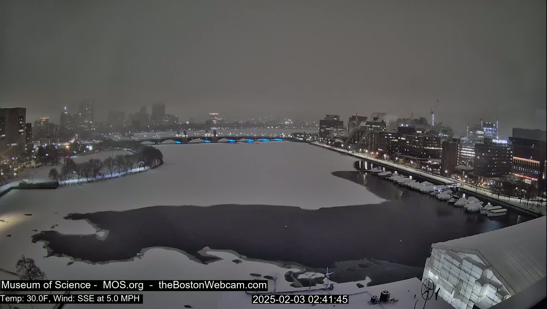 A snow-covered city waterfront at night, with a partially frozen river and boats docked along the shore, under a heavy snow.