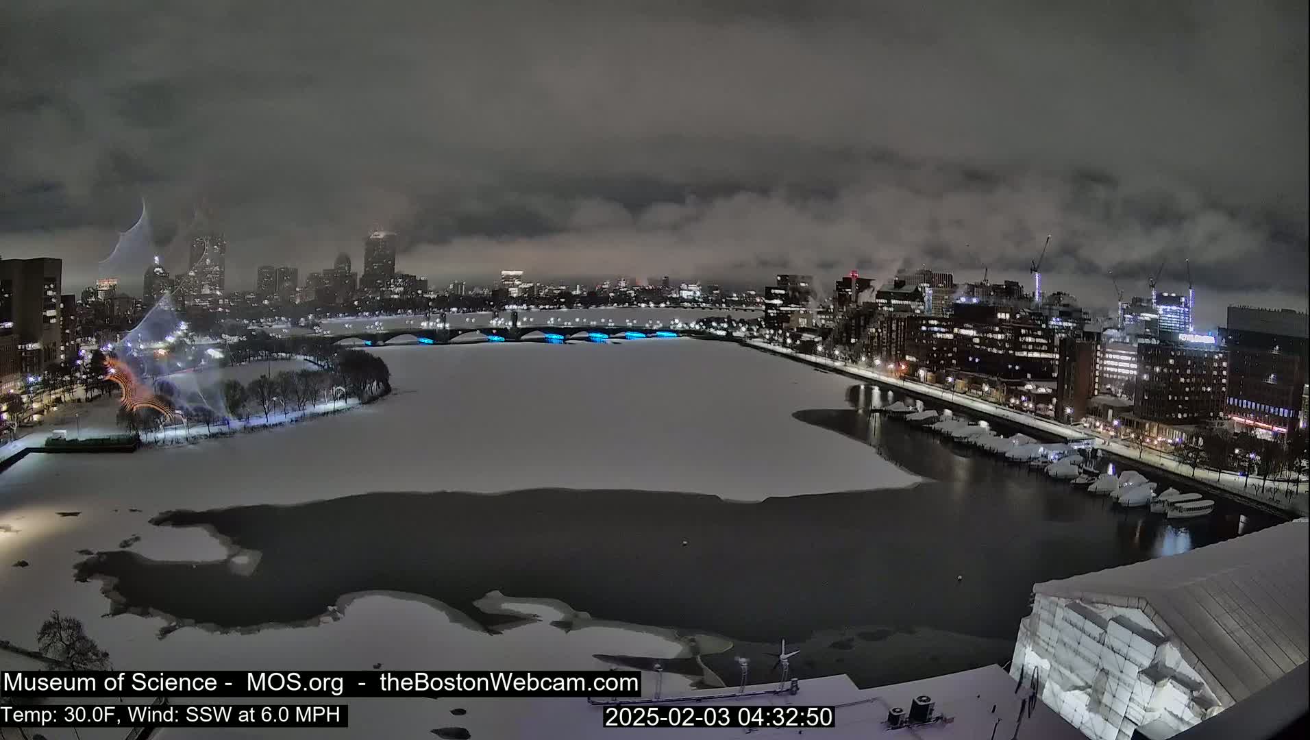A snow-covered city waterfront at night, under a cloudy sky, shows a partially frozen river with illuminated bridges and a cityscape in the background.