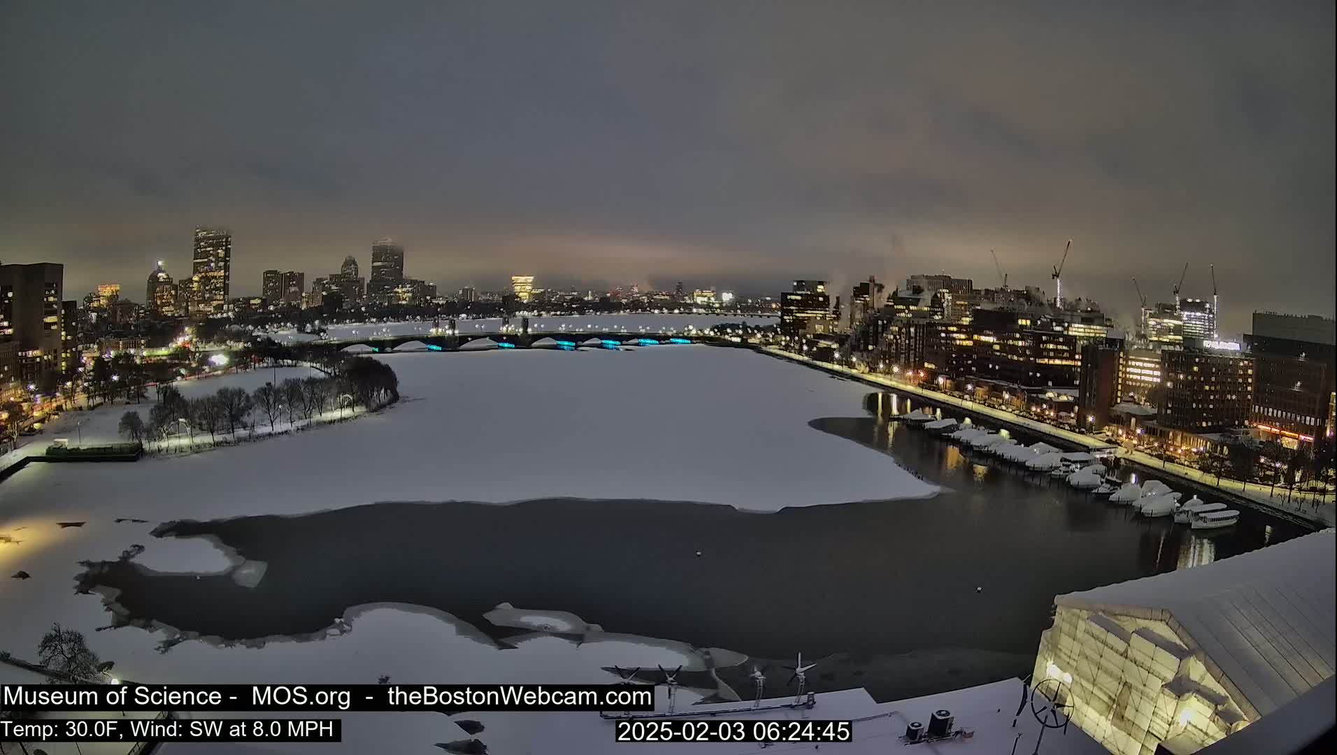 A nighttime, snowy cityscape with a partially frozen river, featuring a bridge and city lights under a dark, overcast sky.