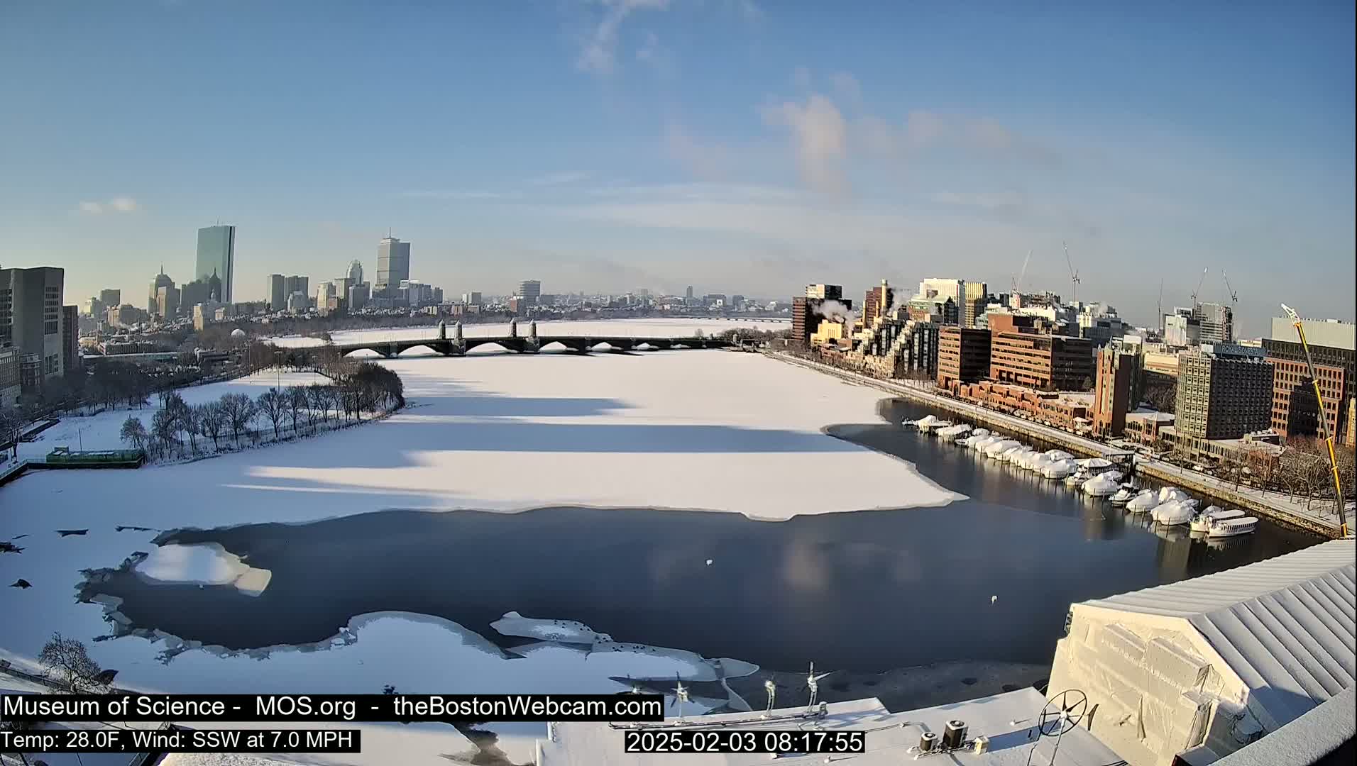 A partly frozen river flows past a snow-covered city, under a clear blue sky.