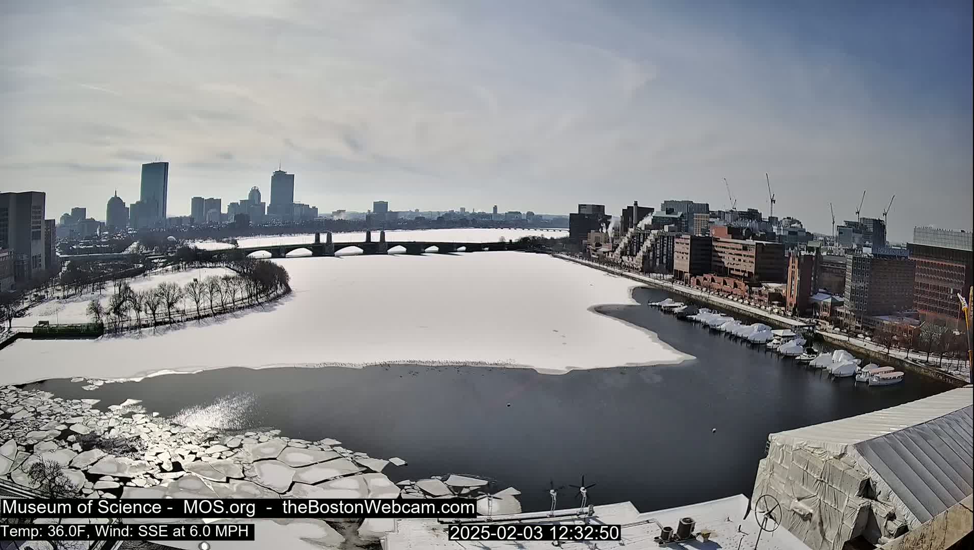 A partly frozen river flows through a city on a sunny, cold day, with snow covering the riverbanks and some buildings in the distance.