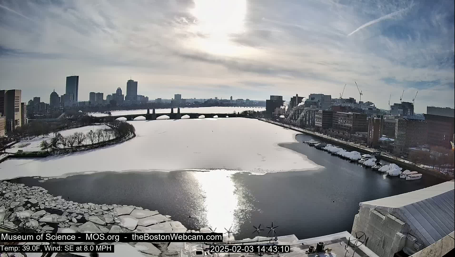 A partly sunny day shows a city skyline across a partially frozen river with ice floes, boats at a marina, and snow on the ground.