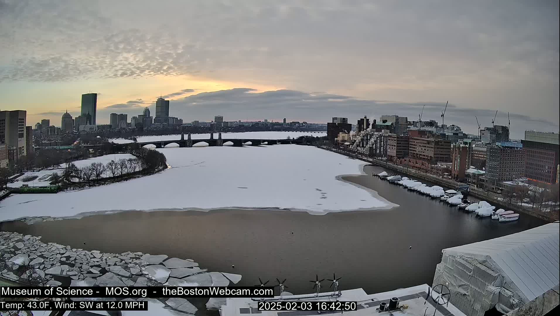 A partly frozen river winds through a snow-covered city landscape under a cloudy, dusky sky.