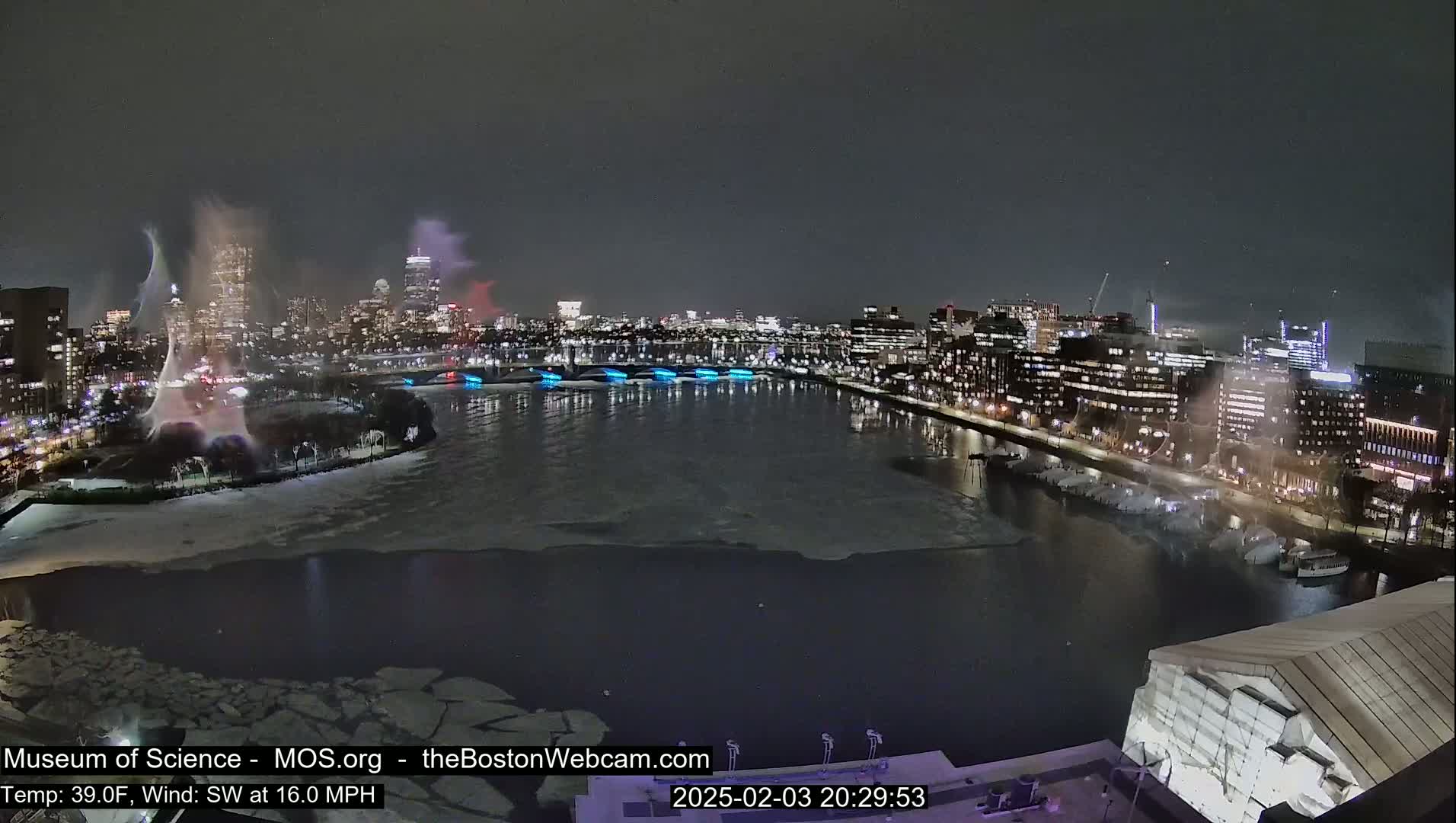 A nighttime city skyline with a partially frozen river, illuminated by city lights and featuring water features, under a dark sky with a southwest wind at 16 mph and a temperature of 39°F.