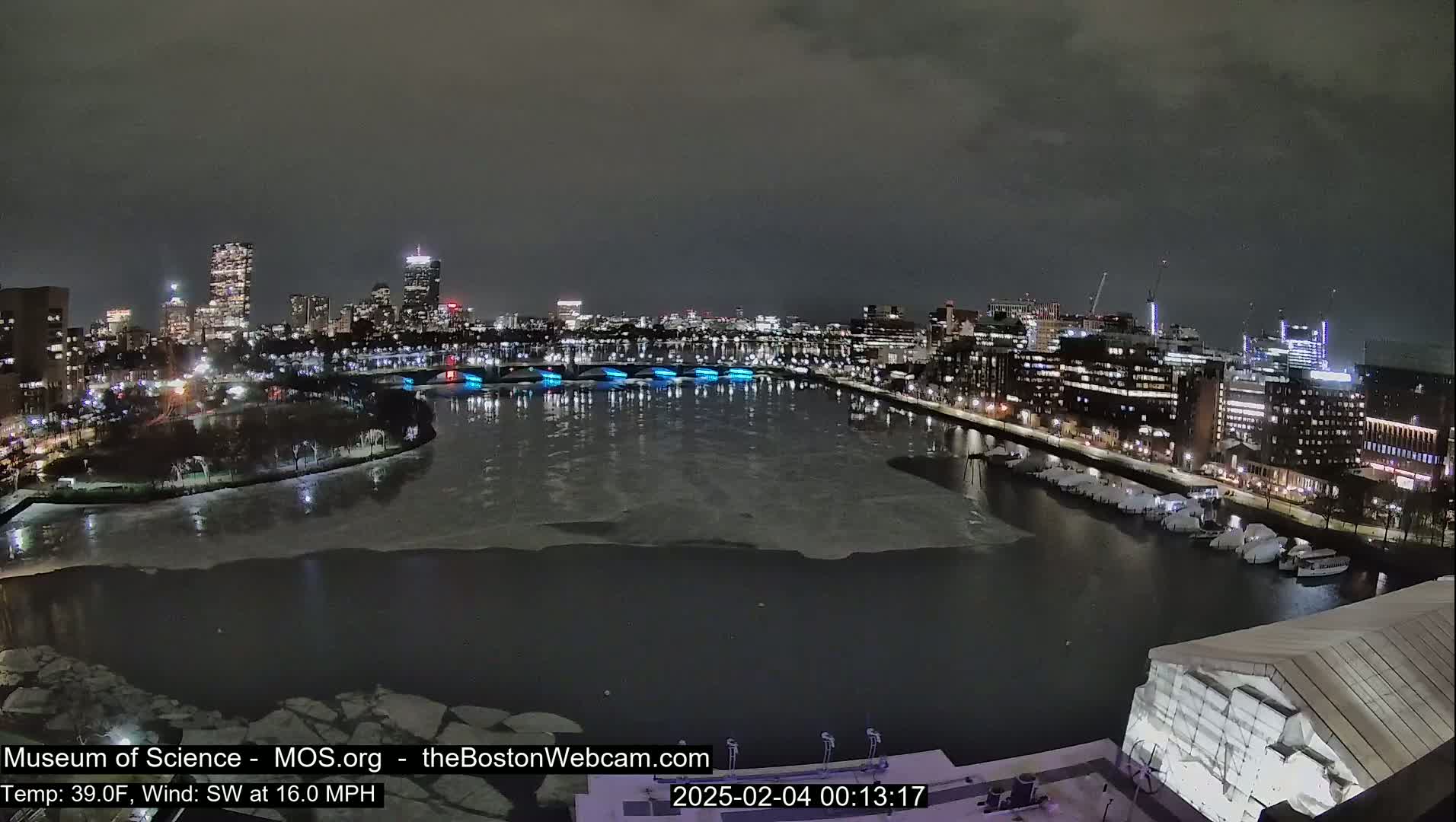 A nighttime cityscape with a partially frozen river, illuminated bridges, and city lights under a dark, overcast sky.