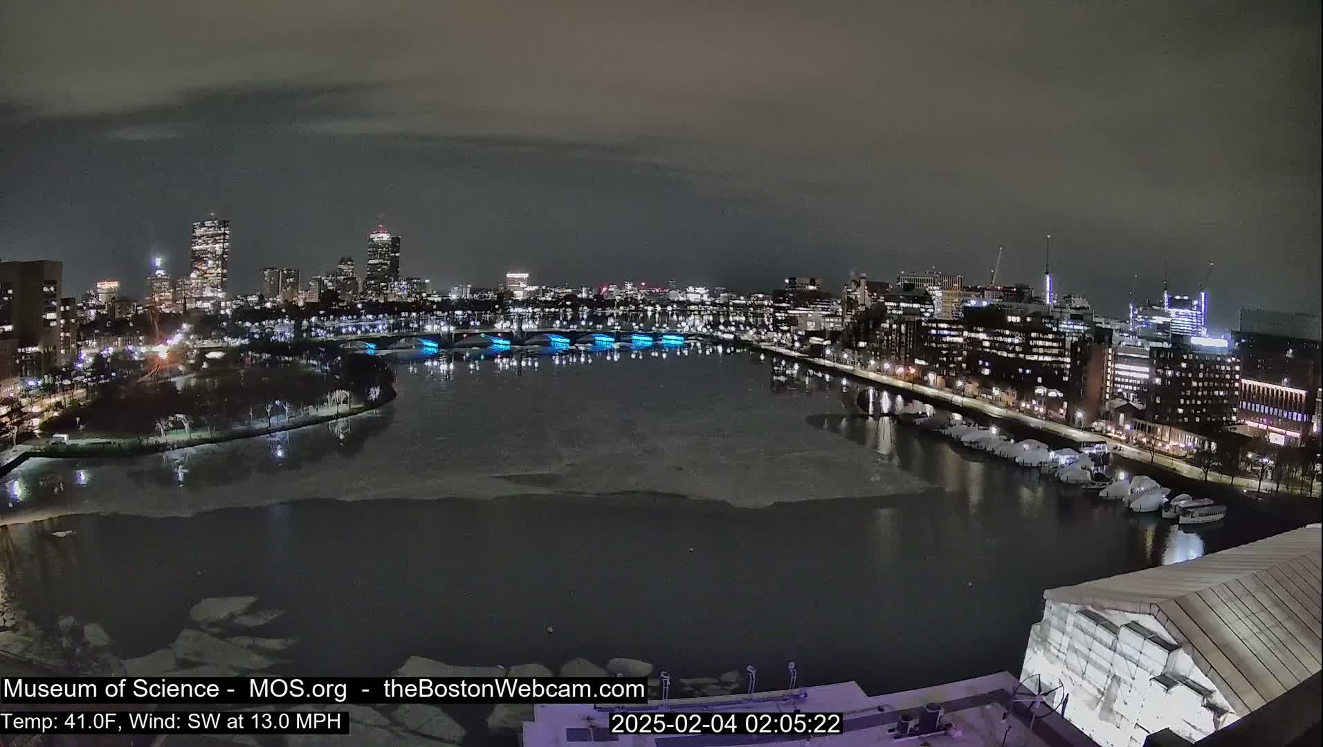 A nighttime view of a city skyline across a partially ice-covered river, under a cloudy sky.