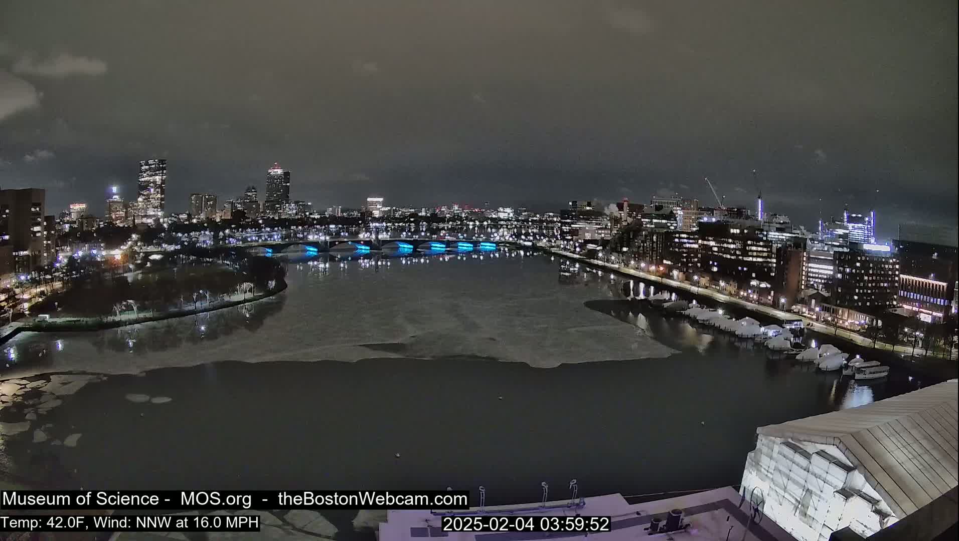 A nighttime cityscape with a partially frozen river, illuminated buildings, and a partly cloudy sky.