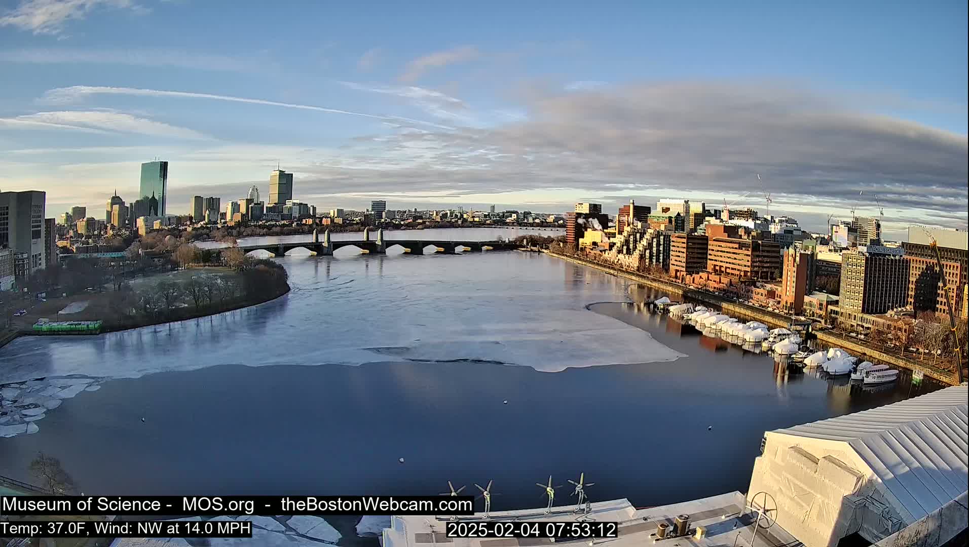 A partially frozen river flows past a city skyline under a partly cloudy sky.