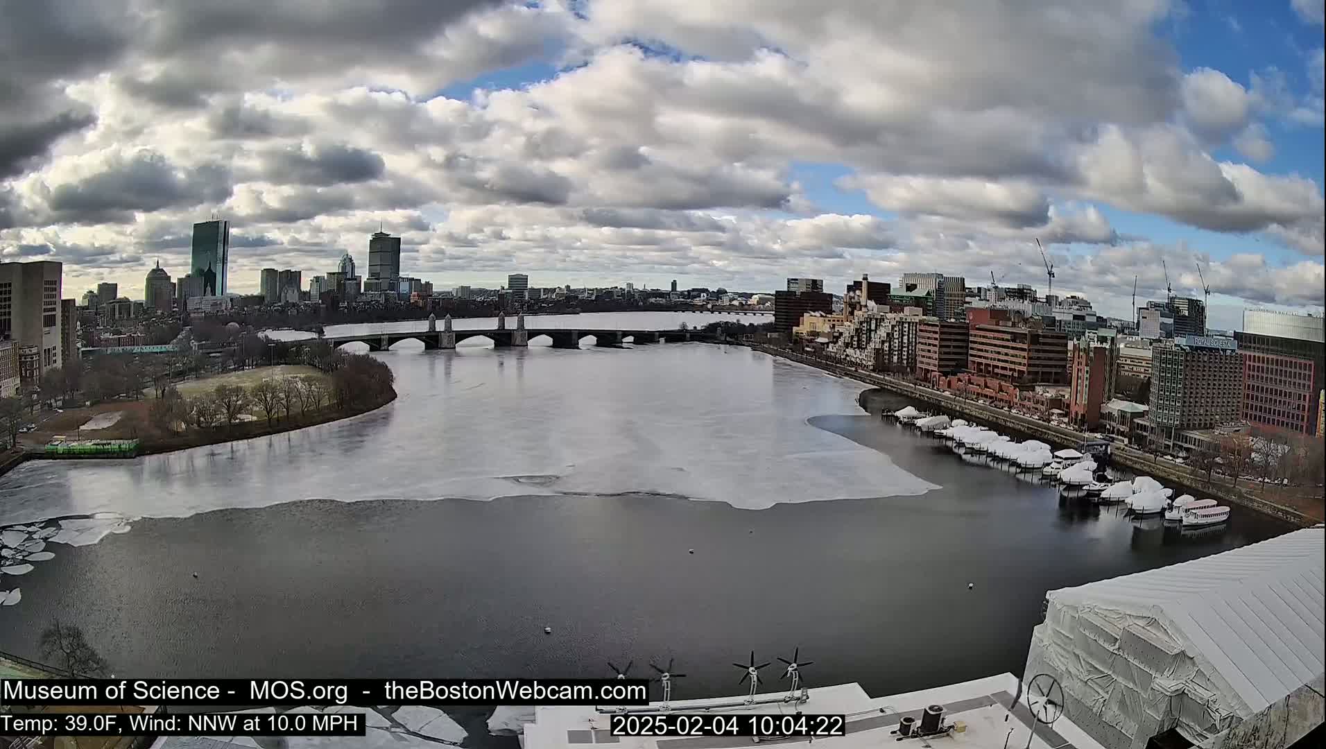 A partly cloudy day shows a partially frozen river flowing past a city skyline with a bridge and docked boats, under a 10 mph wind.
