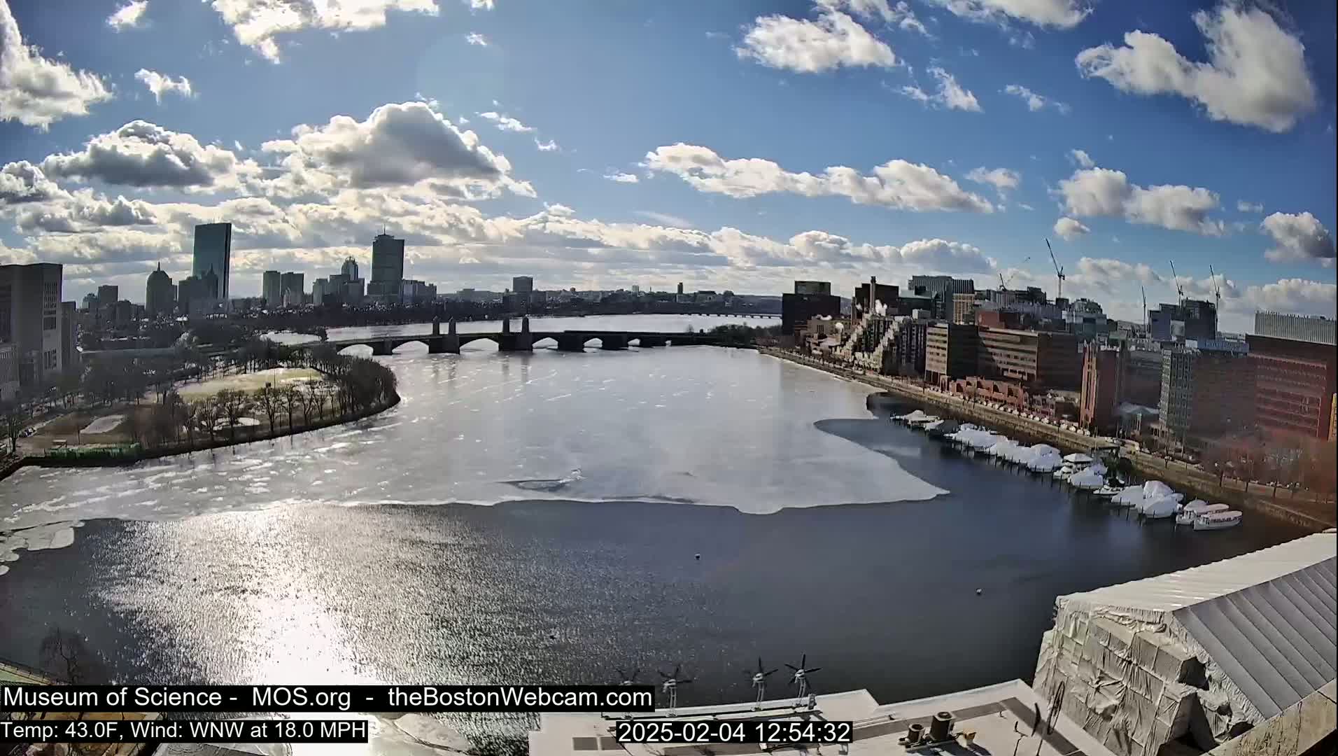 A partly sunny day shows a city skyline across a partially ice-covered river, with boats docked along the shore and construction underway near the camera's location.
