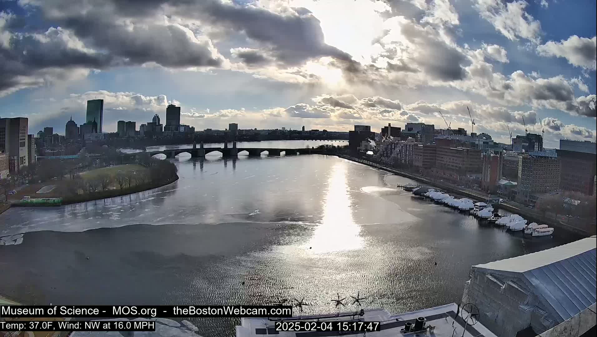 A partly cloudy day with a northwest wind at 16 mph shows a city skyline across a partially frozen river with boats docked along the shore.