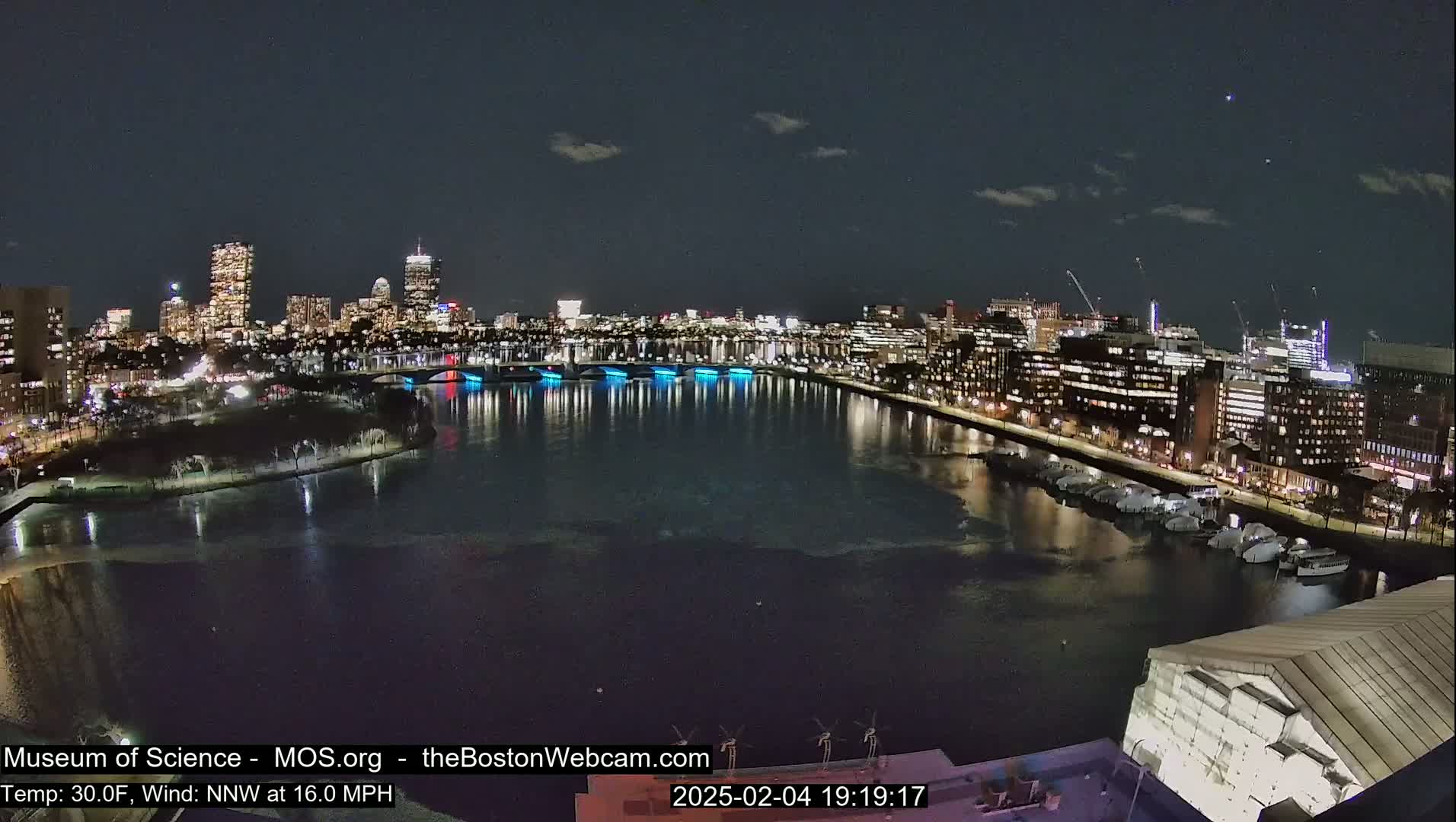 A nighttime view of a city skyline across a body of water, under a partly cloudy sky with a 30°F temperature and 16 mph north-northwest wind.
