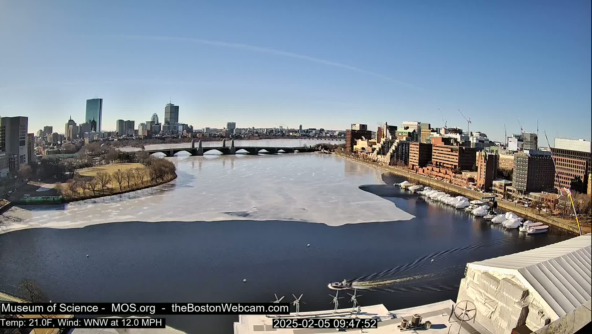 A partly frozen river flows past a city skyline under a clear, sunny sky.