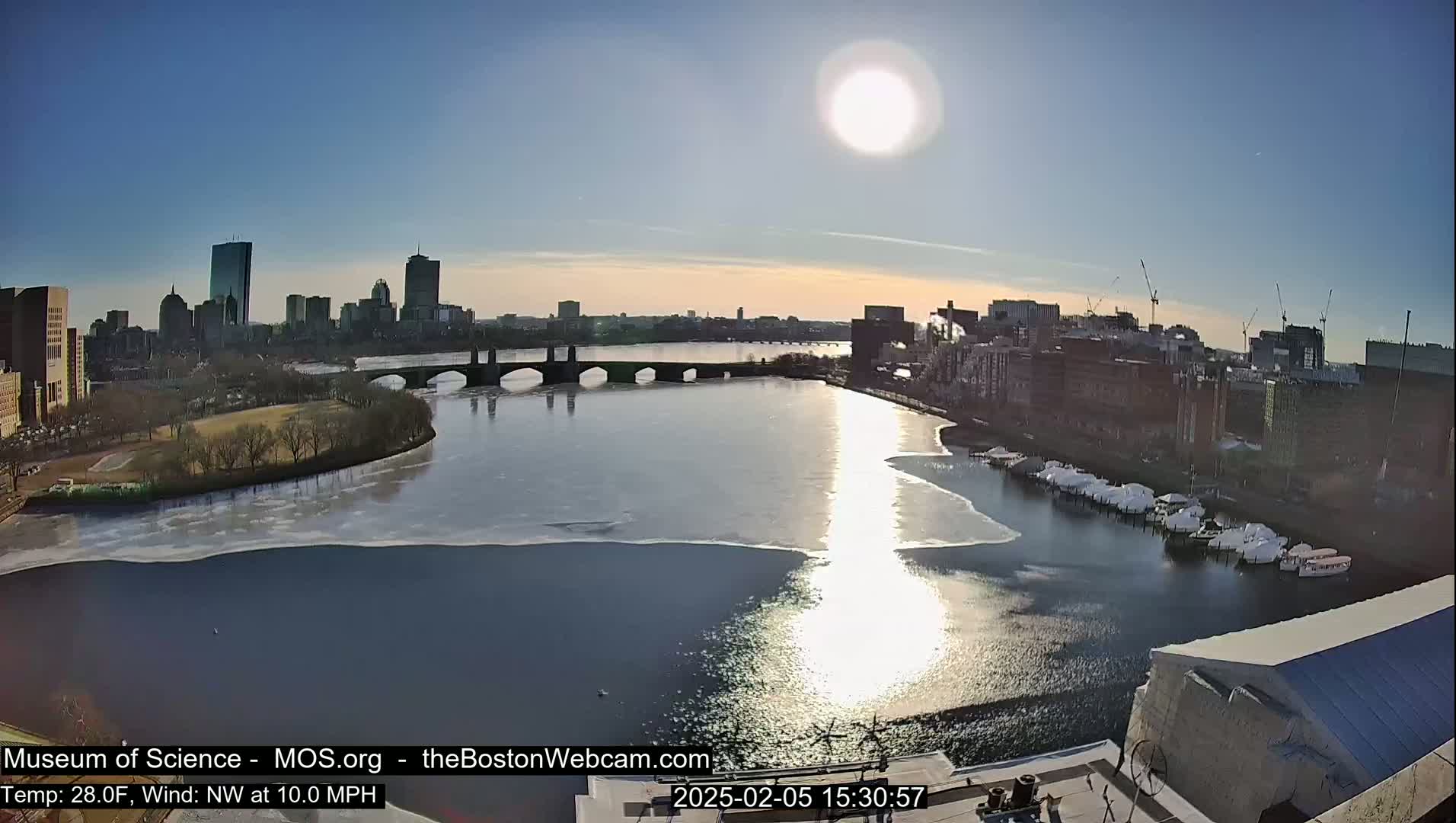 A sunny, cold day shows a partially frozen river flowing past a city skyline and a bridge, with boats docked along the shore.