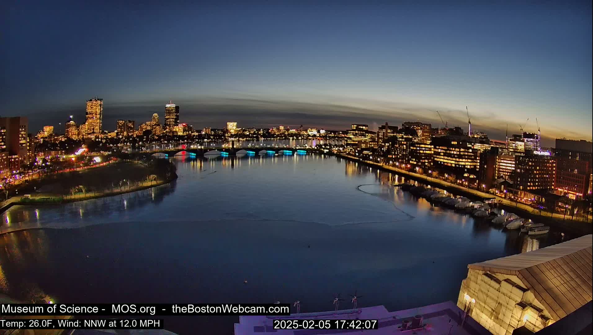 A nighttime cityscape with a river reflecting illuminated buildings and bridges, partially frozen in the cold, under a twilight sky.