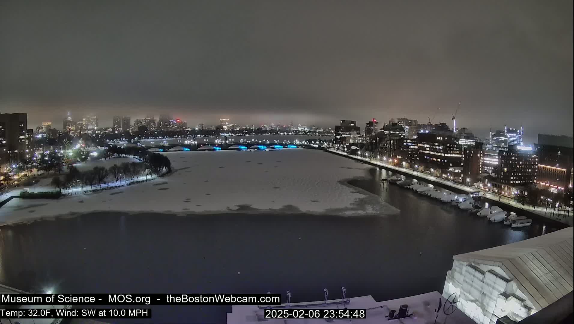 A nighttime view of a snow-covered city waterfront, with a partially frozen river and a cityscape in the background under hazy conditions.