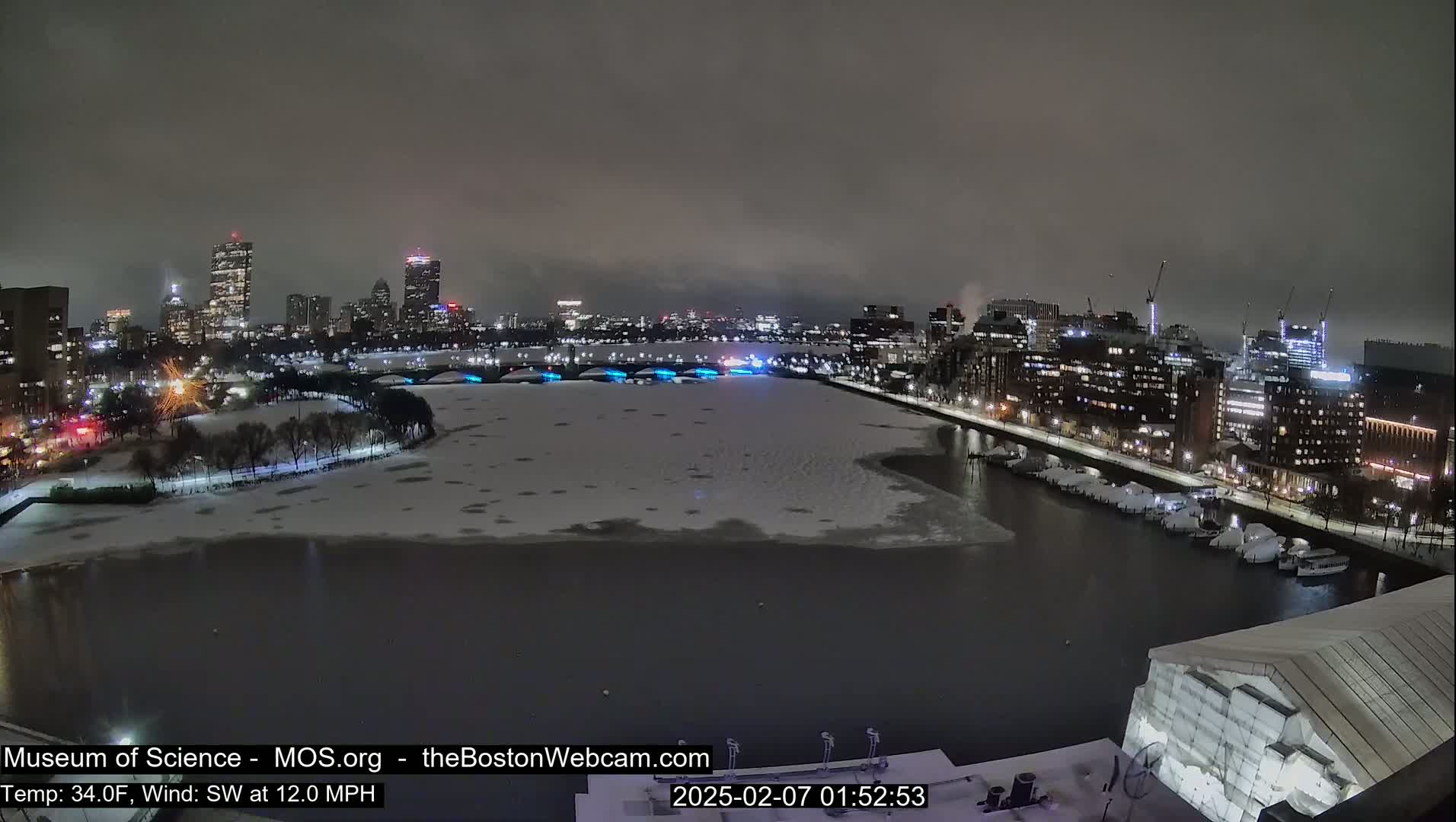 A nighttime view of a snow-covered river and city skyline under a dark, overcast sky.