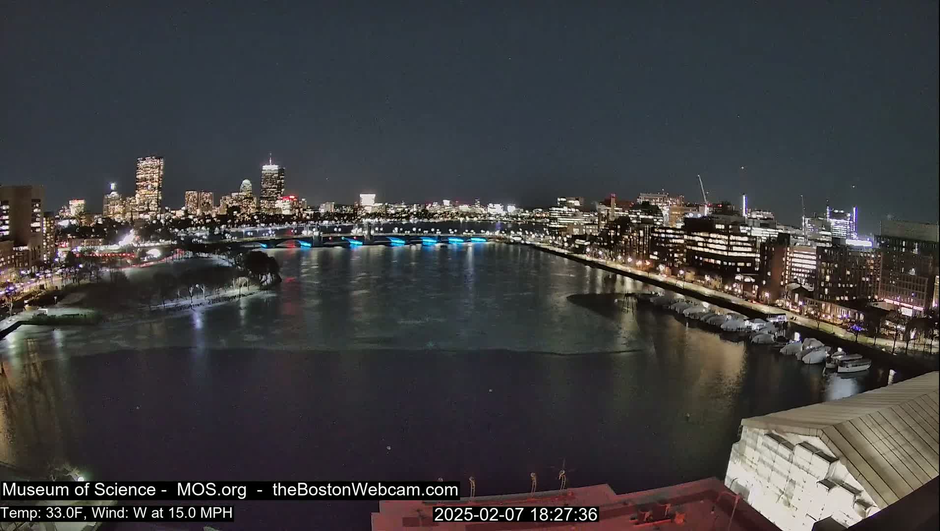 A nighttime view of a city skyline across a partially frozen river, under a clear sky.