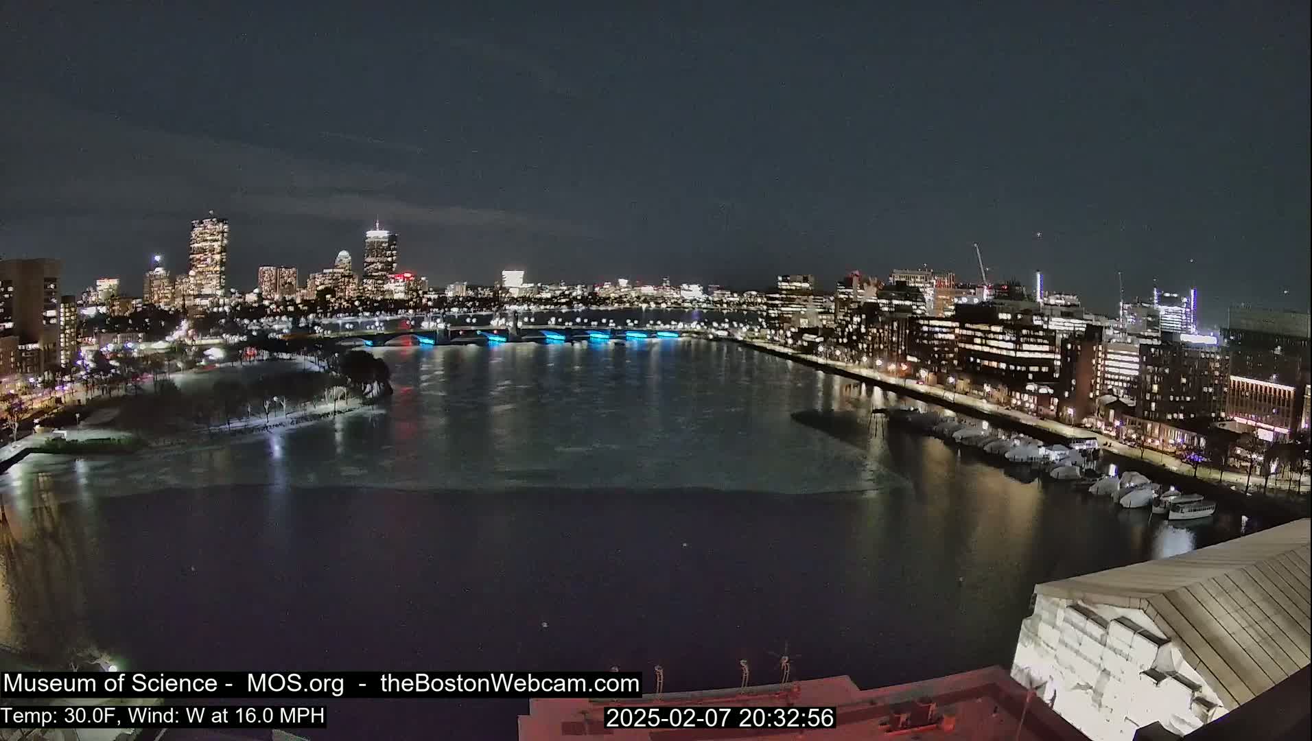 A nighttime view of a city skyline across a partially frozen river, with a 30°F temperature and a 16 mph west wind.