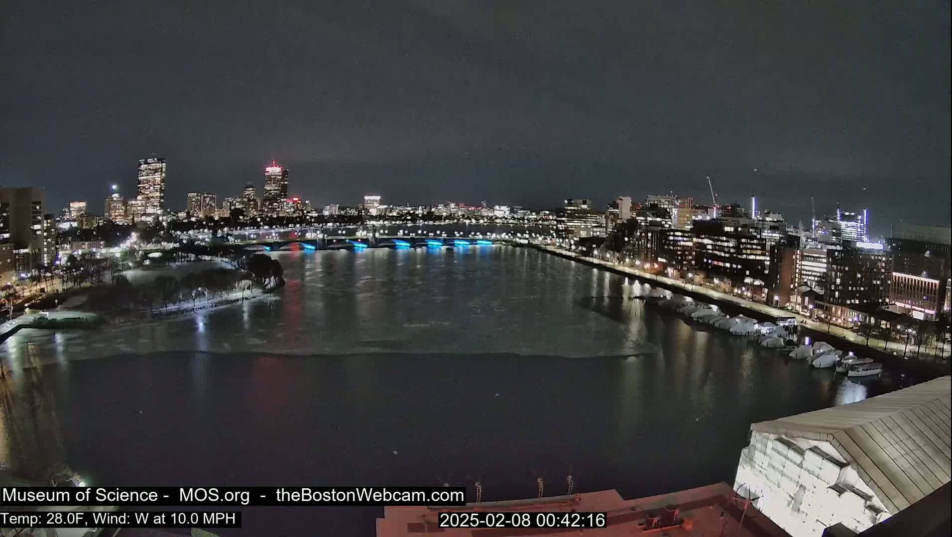 A nighttime cityscape with a partially frozen river, illuminated bridges, and docked boats under a clear, dark sky.