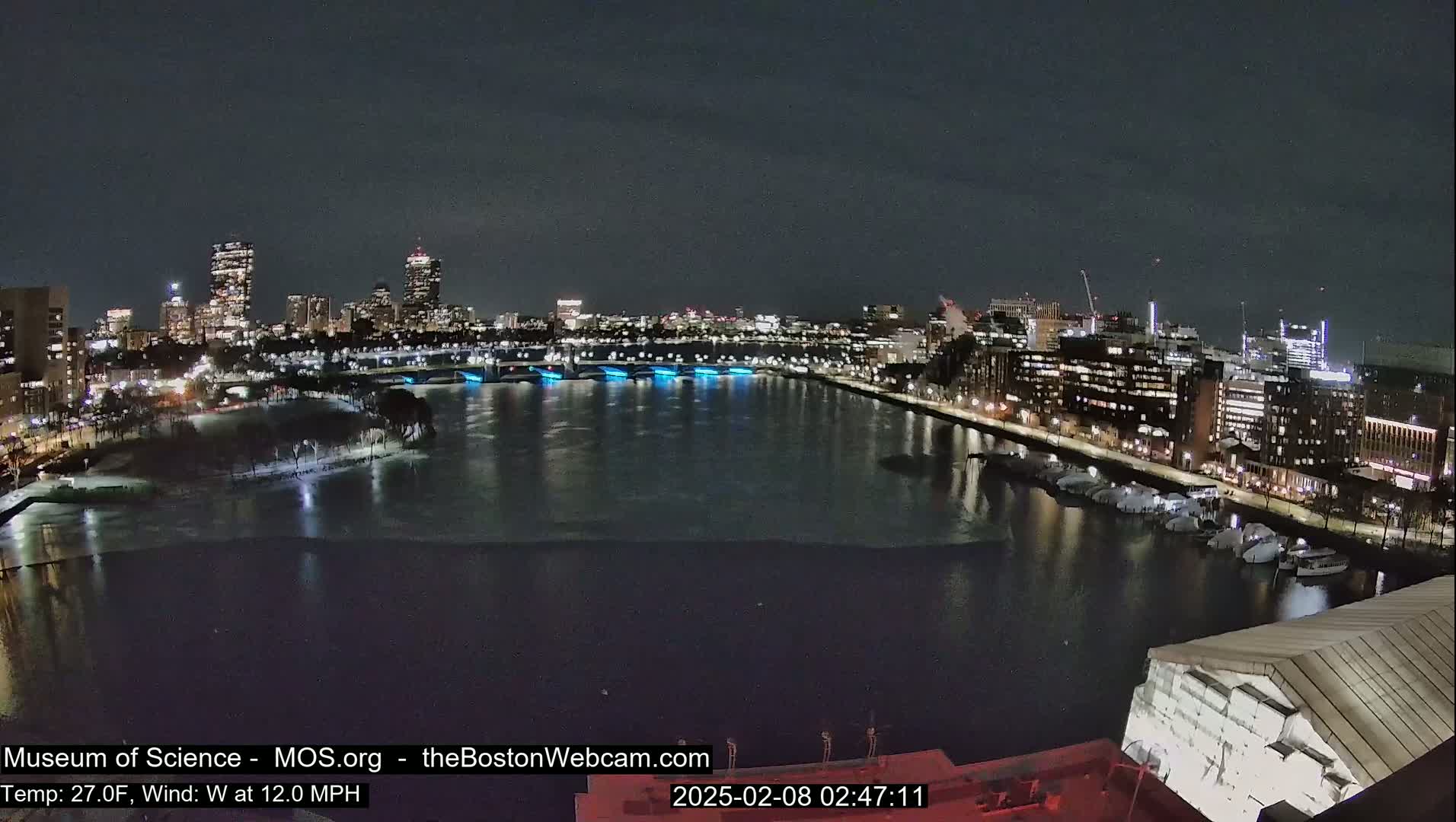 A nighttime cityscape with a partially frozen river, illuminated buildings, and a  wind of 12 mph at a temperature of 27°F.