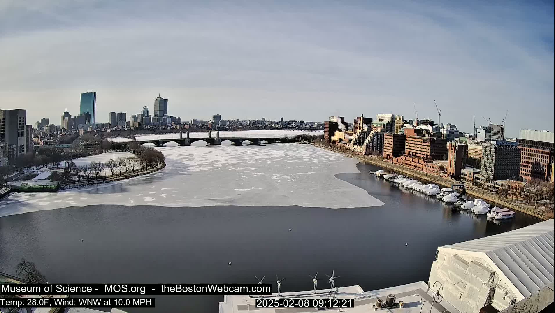 A partially frozen river flows past a city skyline under a clear, sunny sky.