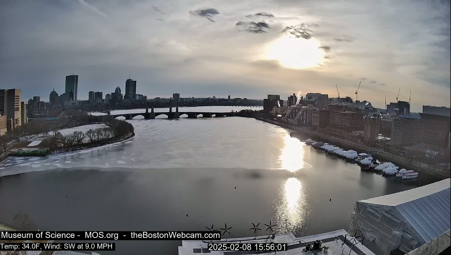 A partially frozen river flows past a city skyline under a partly cloudy sky, with the sun setting.
