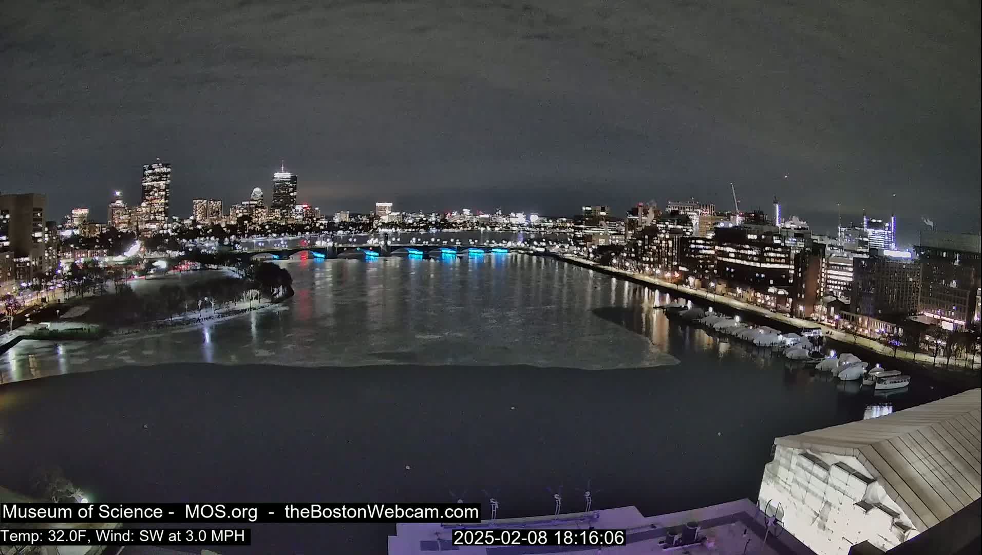 A nighttime cityscape with a partially ice-covered river, illuminated bridges, and a city skyline under a dark sky.