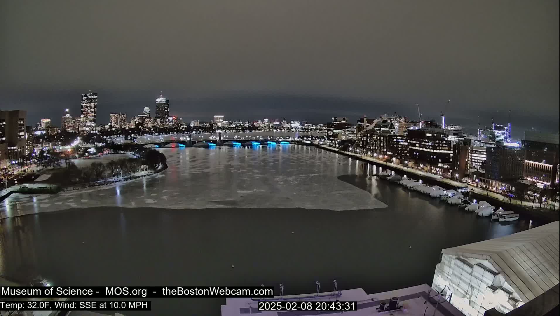 A nighttime view of a partially frozen river with a cityscape in the background under a dark sky.