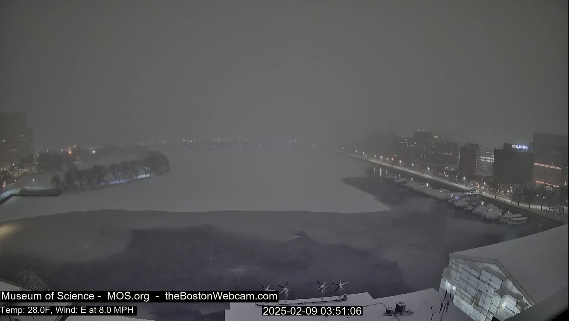 A snow-covered waterfront scene at night, with buildings and docked boats visible through heavy snowfall.