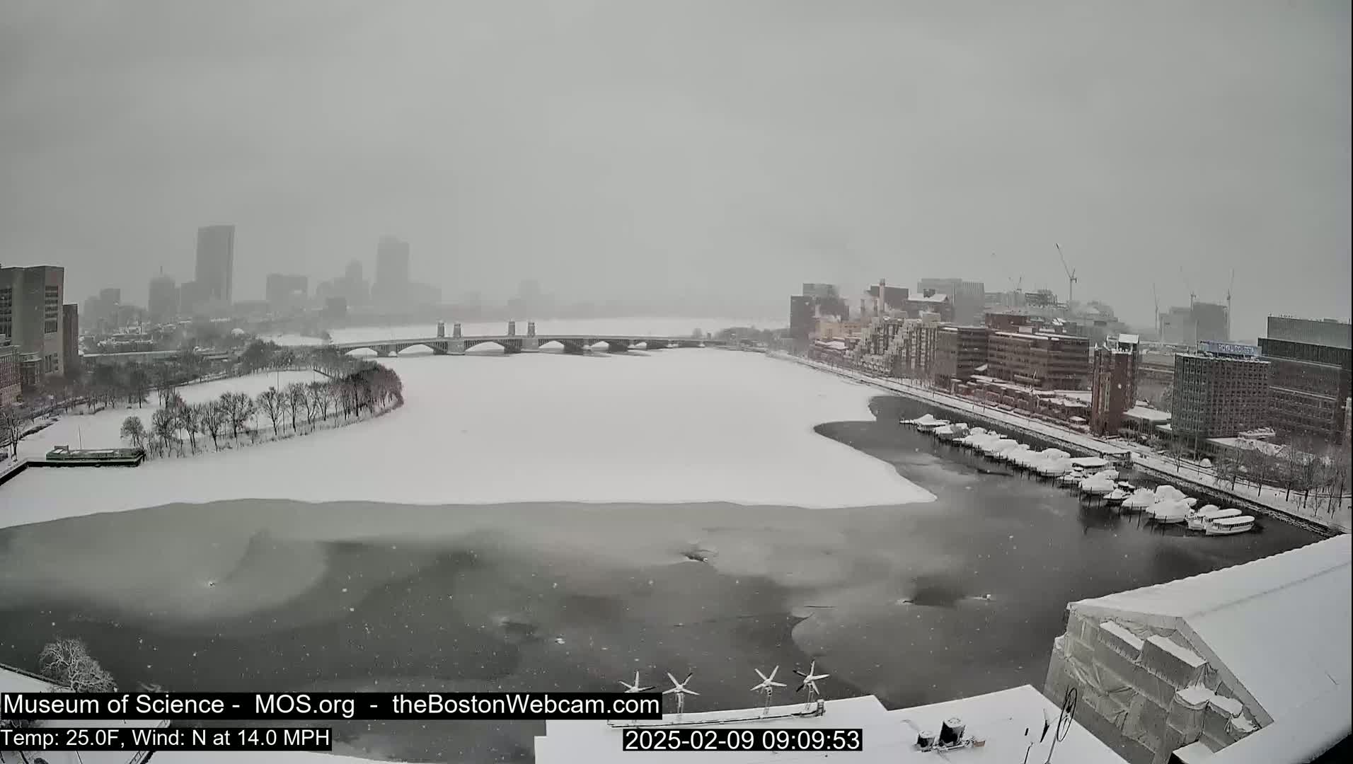 A snow-covered cityscape with a river partially frozen over, viewed from a high vantage point during a snowstorm.
