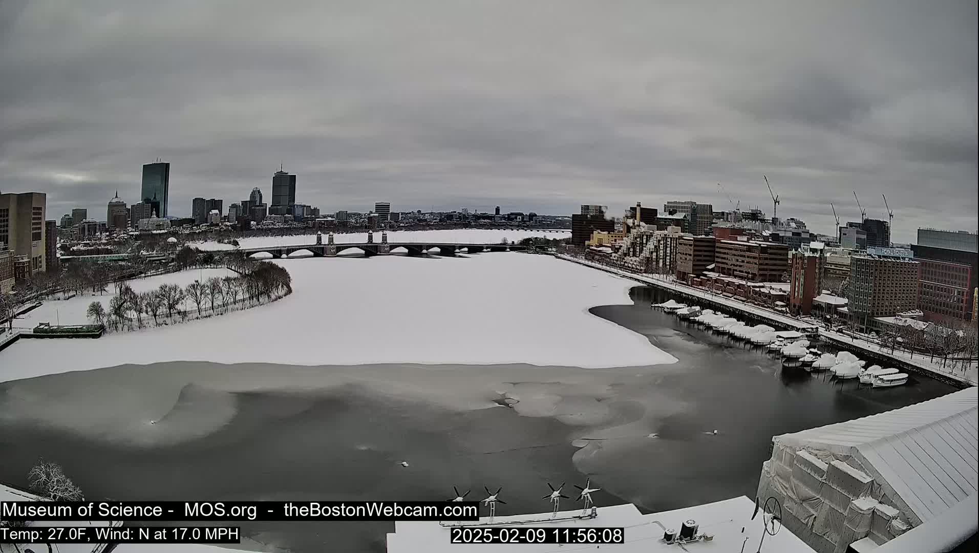 A snow-covered city skyline is visible across a partially frozen river, under an overcast sky.