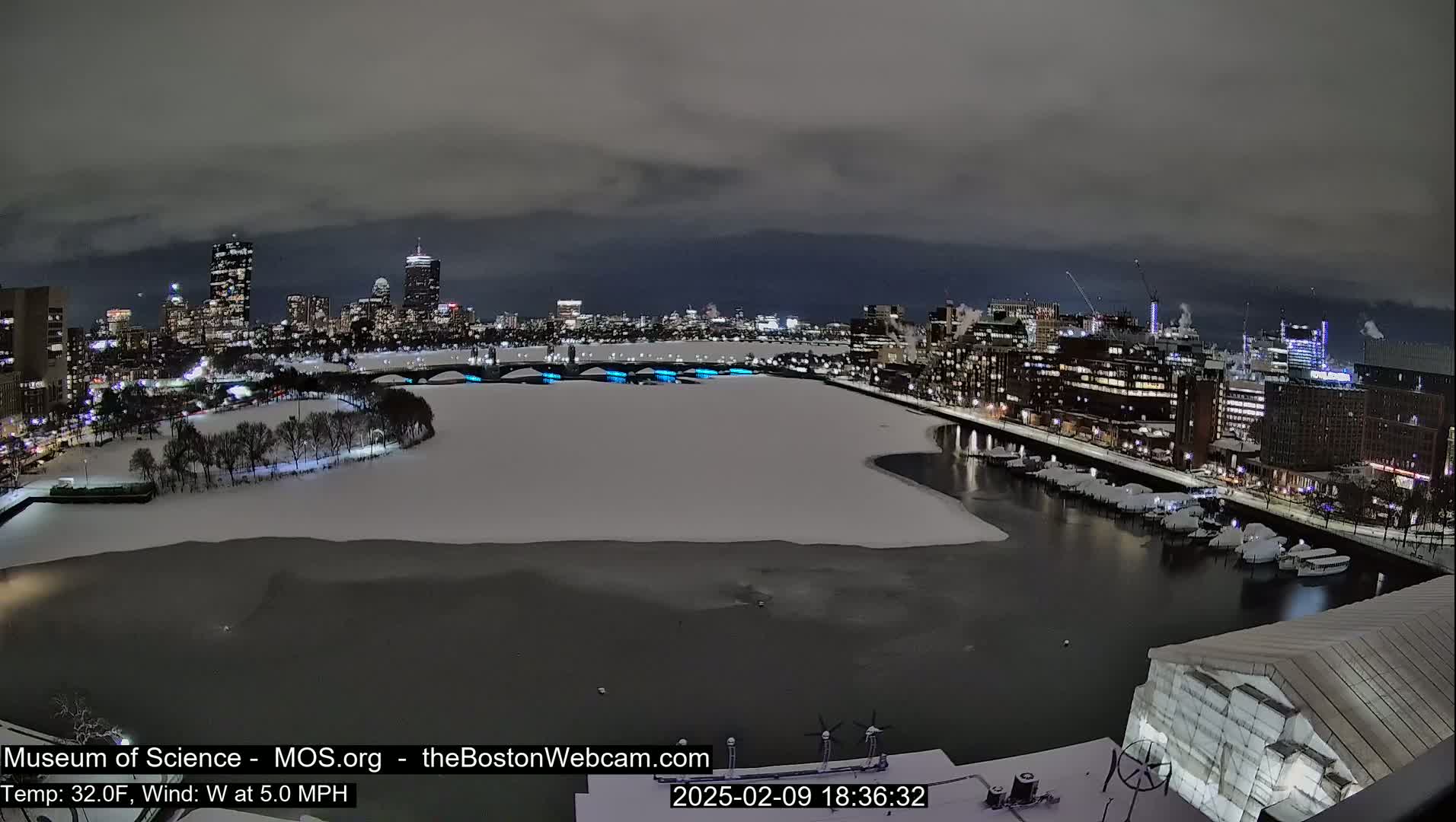 A snow-covered city waterfront at night, with a partially frozen river and illuminated bridges, under a cloudy sky.