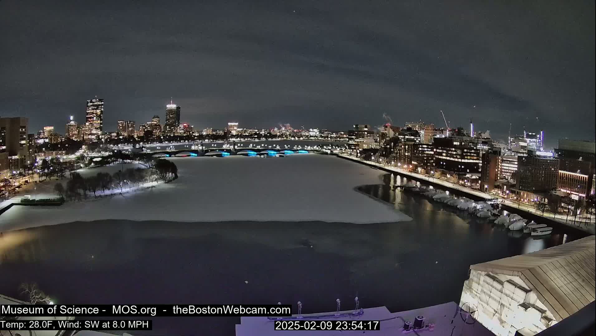 A nighttime view of a snow-covered city waterfront with a partially frozen river, illuminated buildings, and bridges.