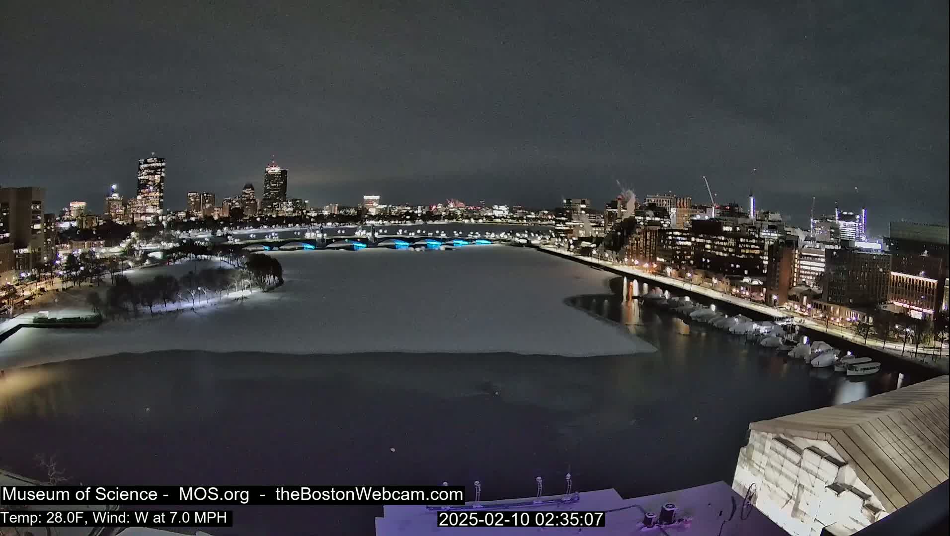 A nighttime cityscape with a partially frozen river, snow-covered ground, and city lights reflecting on the water under a dark sky.