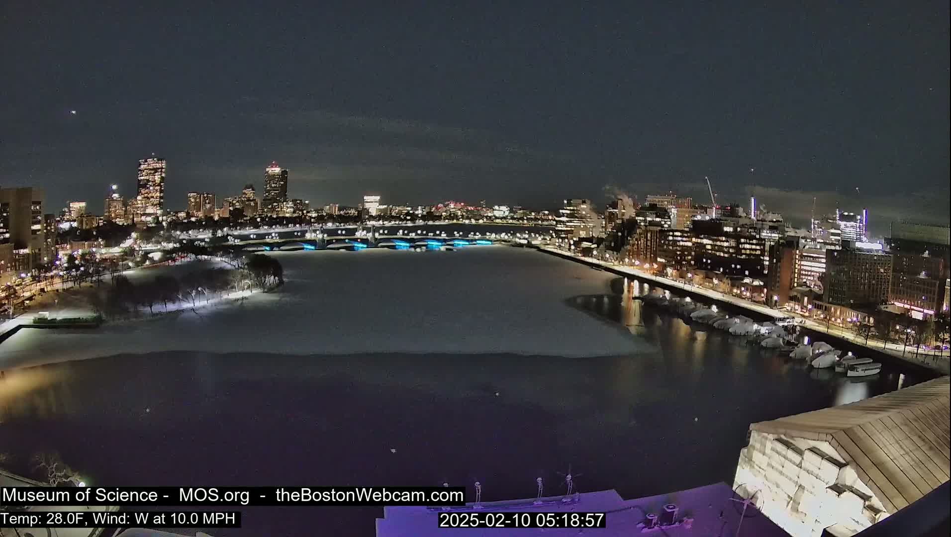 A nighttime cityscape with a partially frozen river, illuminated buildings, and a snow-covered shoreline under a clear, dark sky.
