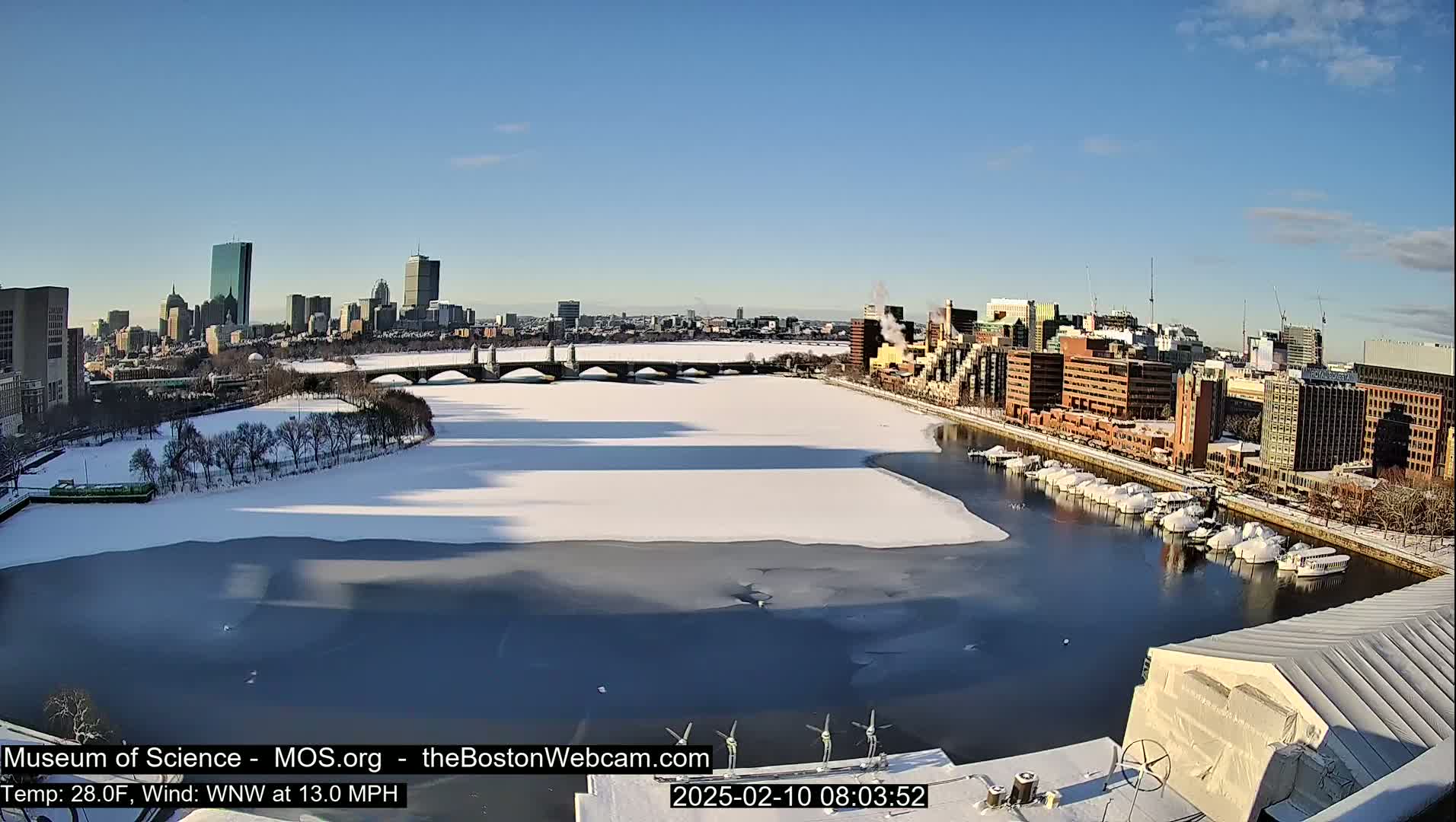 A partially frozen river flows past a snow-covered city, under a clear blue sky.