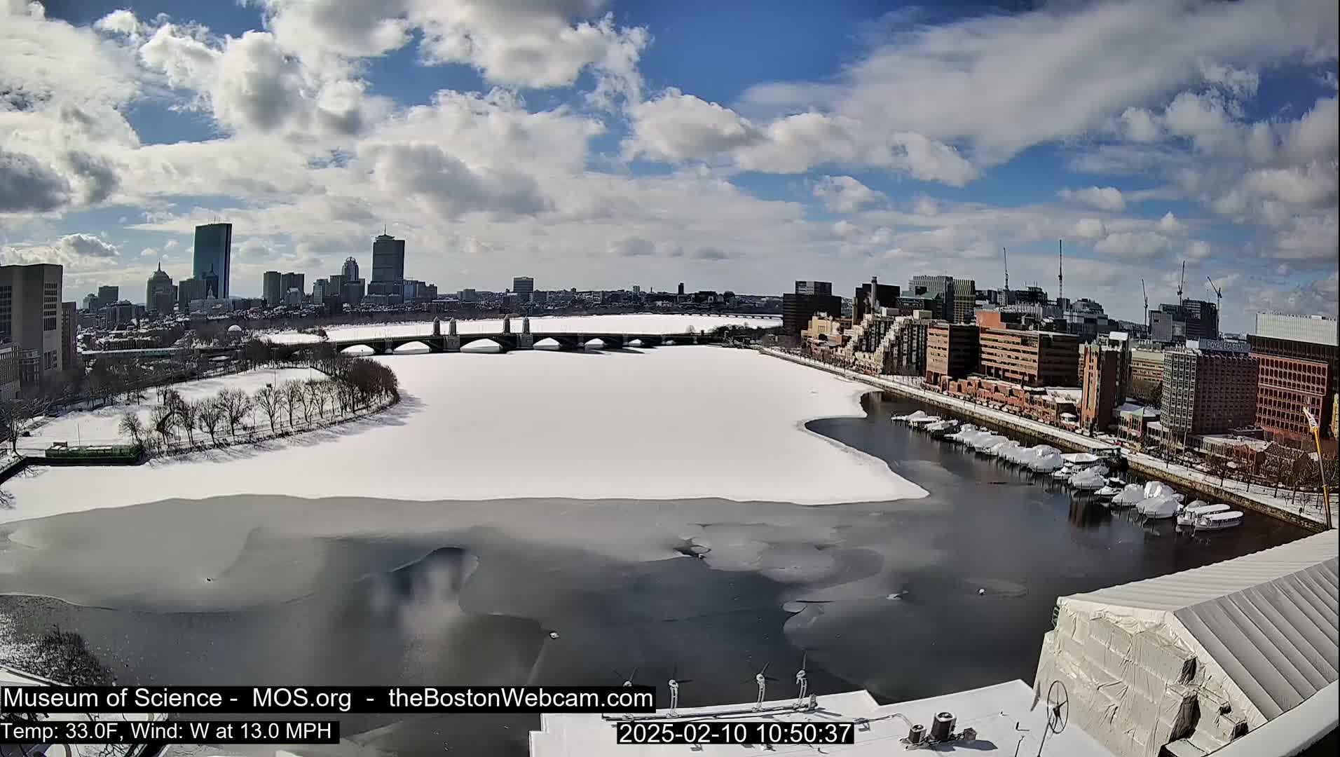A partially frozen river flows past a snow-covered city, under a partly sunny sky.