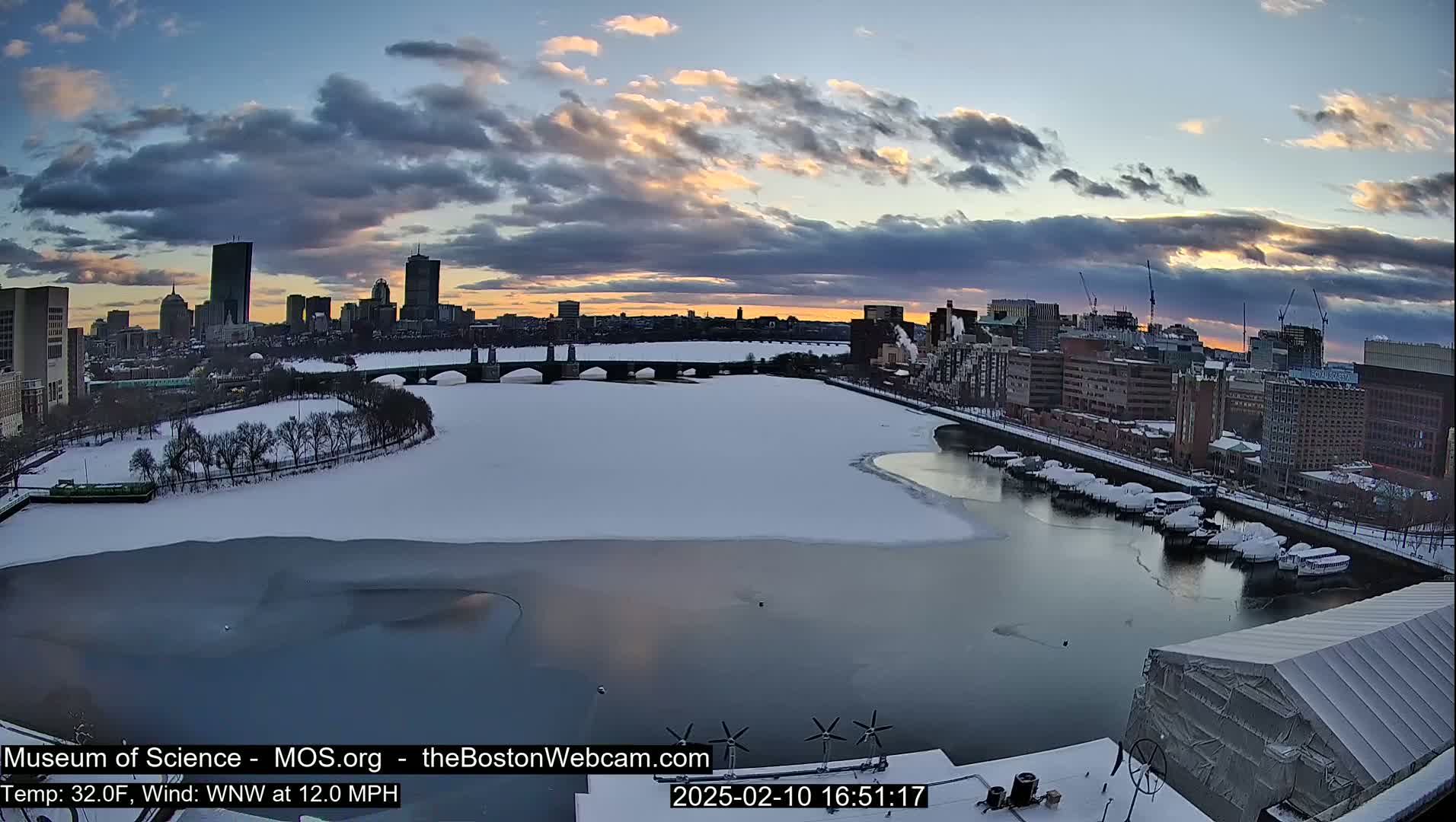 A snow-covered cityscape and river, partially frozen, are viewed at sunset under a cloudy sky.