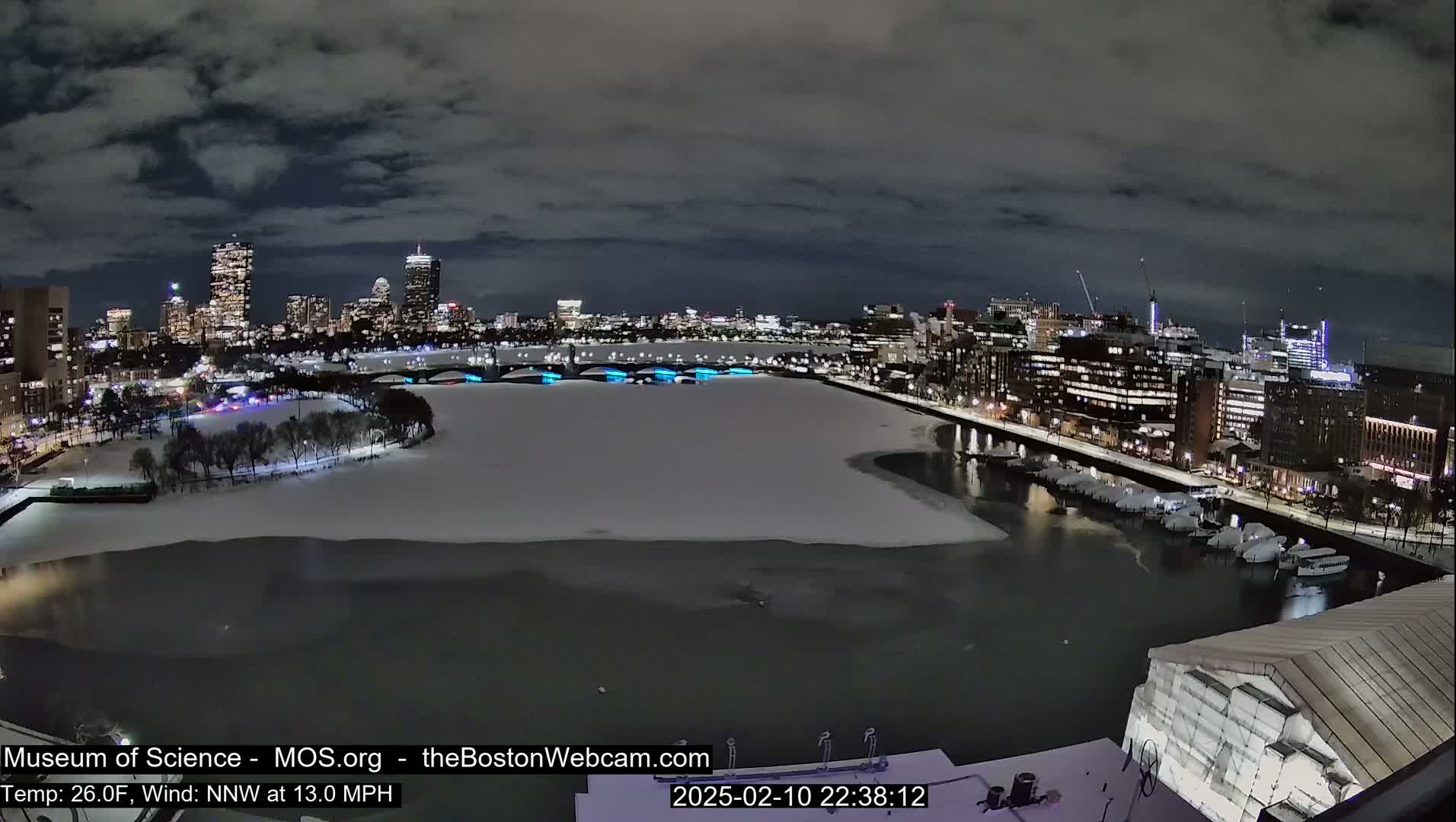 A nighttime view of a snow-covered city waterfront, with a partially frozen river and a cityscape illuminated in the background under a cloudy sky.