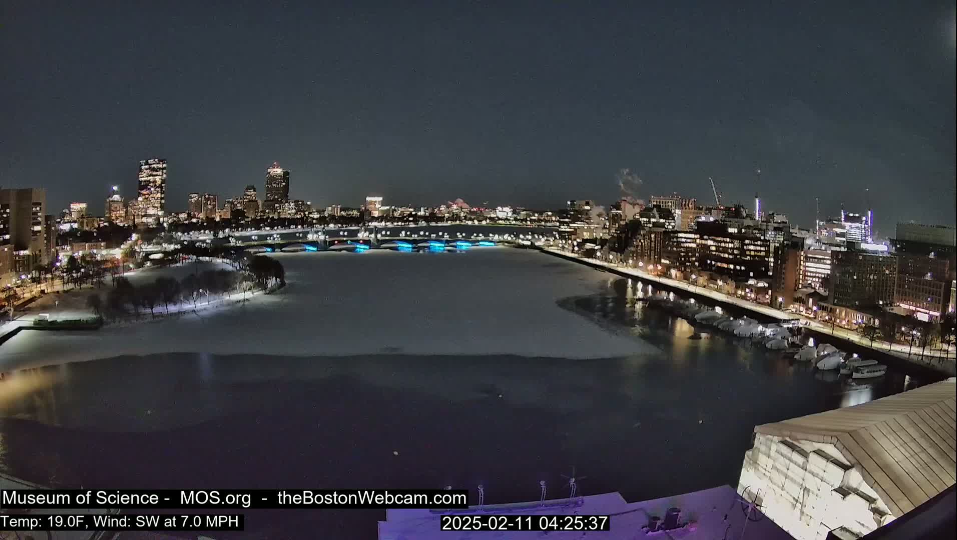 A nighttime view of a snow-covered city waterfront with a partially frozen river, under a clear, dark sky.
