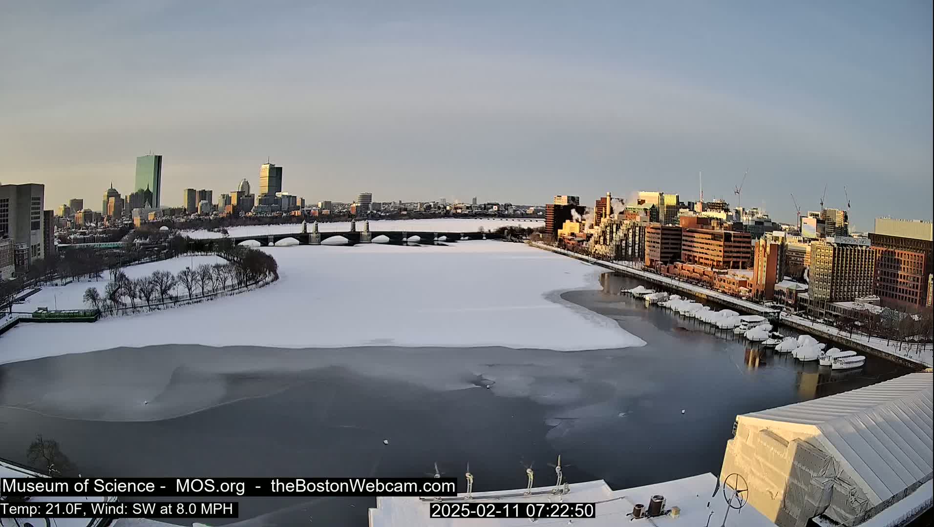 A partly frozen river flows past a snow-covered city, with a bridge and numerous buildings visible under a clear, sunrise sky.